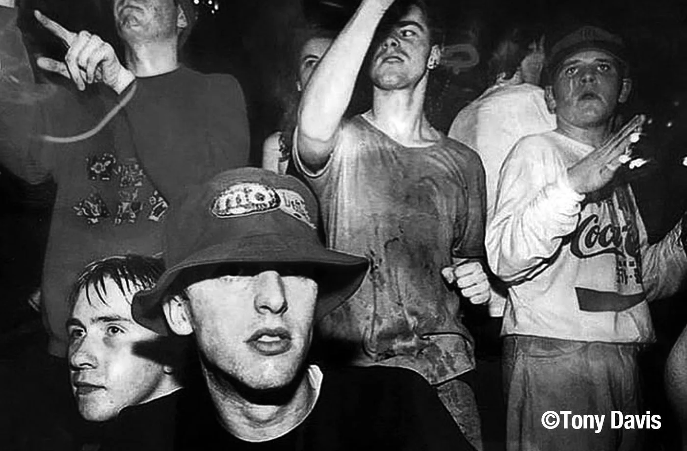 Black and white photo of a group of young people at a concert or party, some with raised hands and attentive expressions, one person with a bucket hat and sunglasses, casual clothing and a Coca-Cola shirt visible.