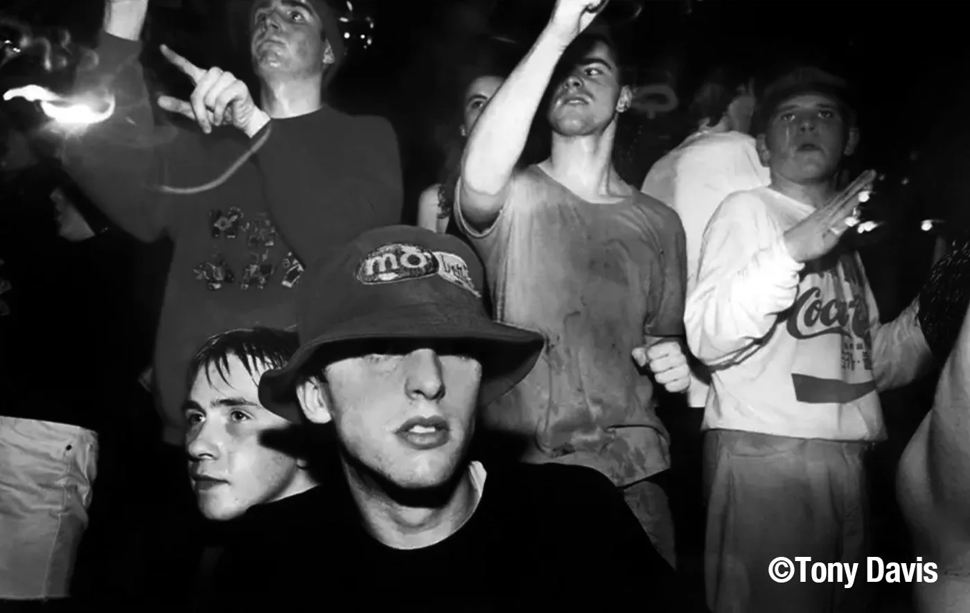 Group of young men at a party or concert, some are singing or dancing, in a black and white photo with a copyright watermark 'Tony Davis'.
