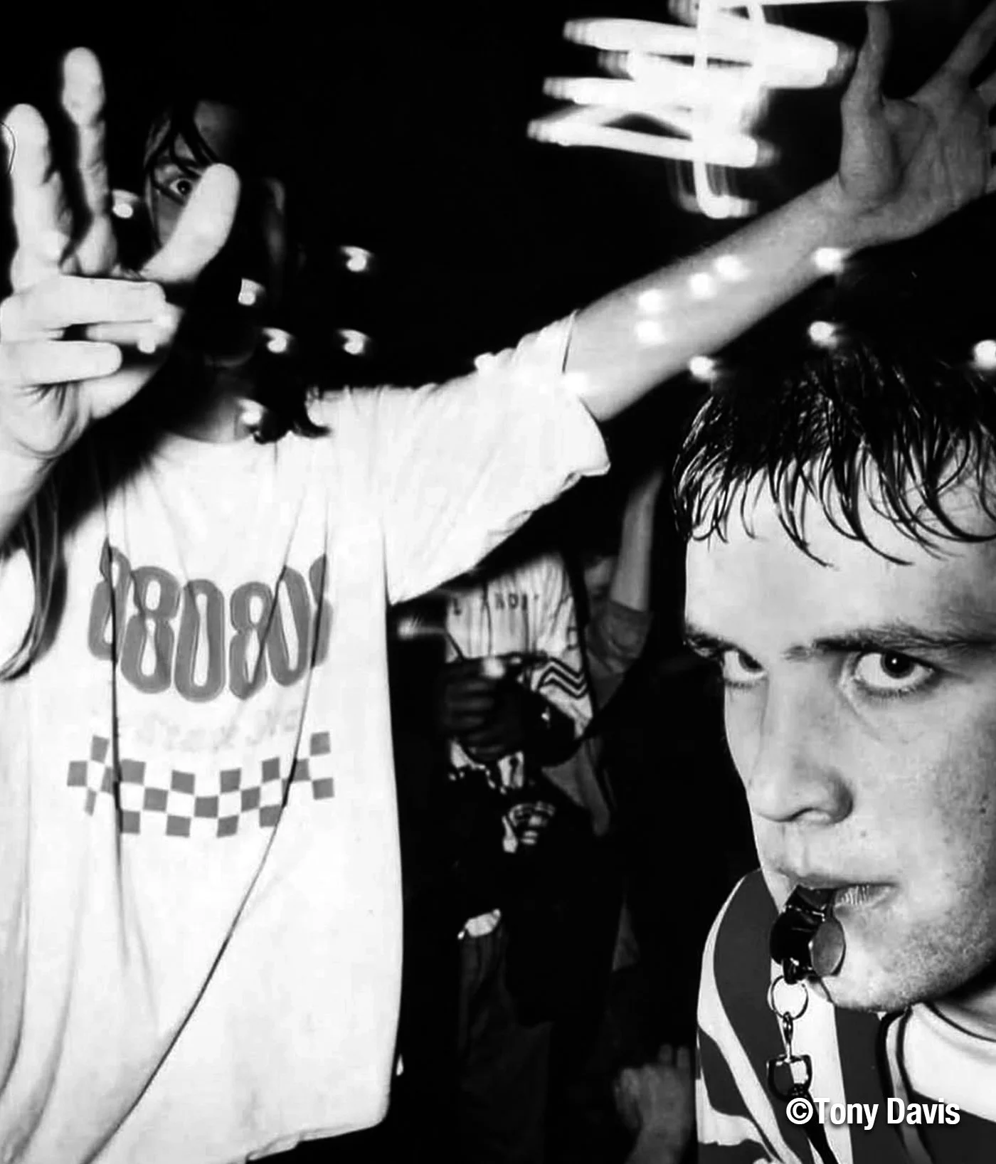 Two young men are closely positioned, one looking directly at the camera with a serious expression, and the other partially obscured, raising his hand with fingers splayed. The scene appears to be in a dark setting with some light reflections.