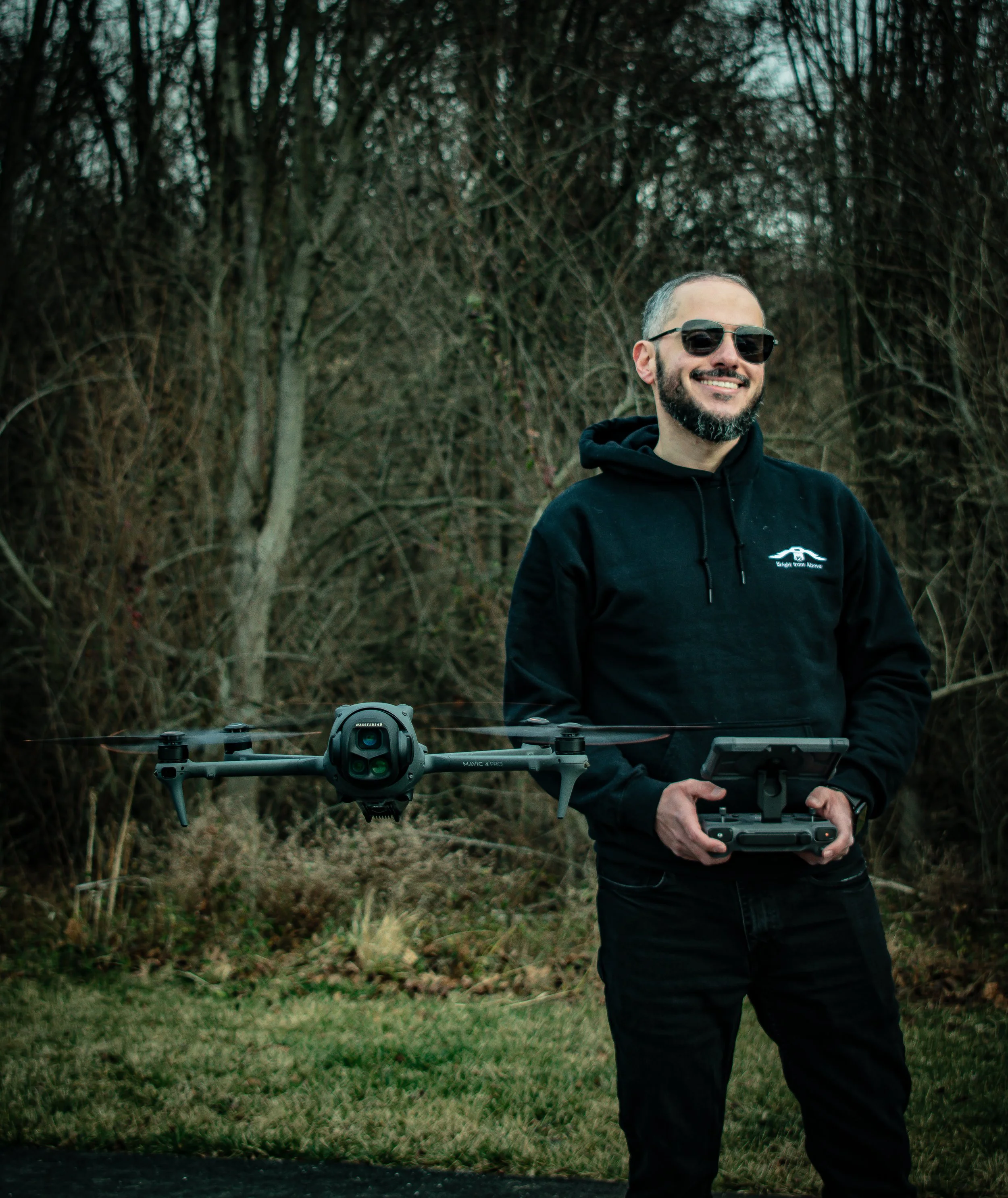 Man smiling outdoors holding a drone with a controller in front of a wooded background. Showcasing DJI Mavic 4 Pro used for corporate and event photography, as well as portraits with it's power camera setup.