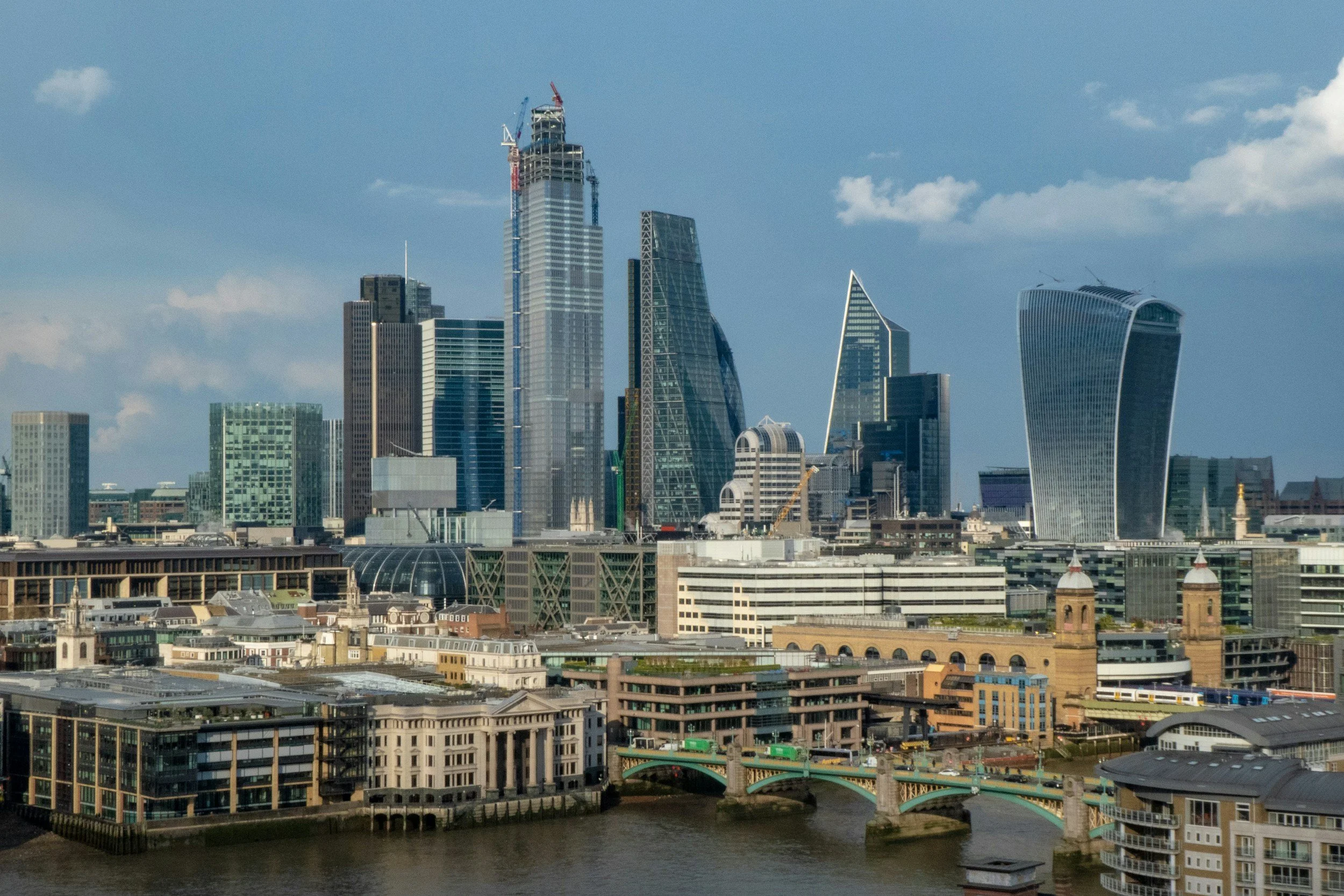Skyline of a modern city with tall skyscrapers, including a building under construction, and a river in the foreground with bridges.