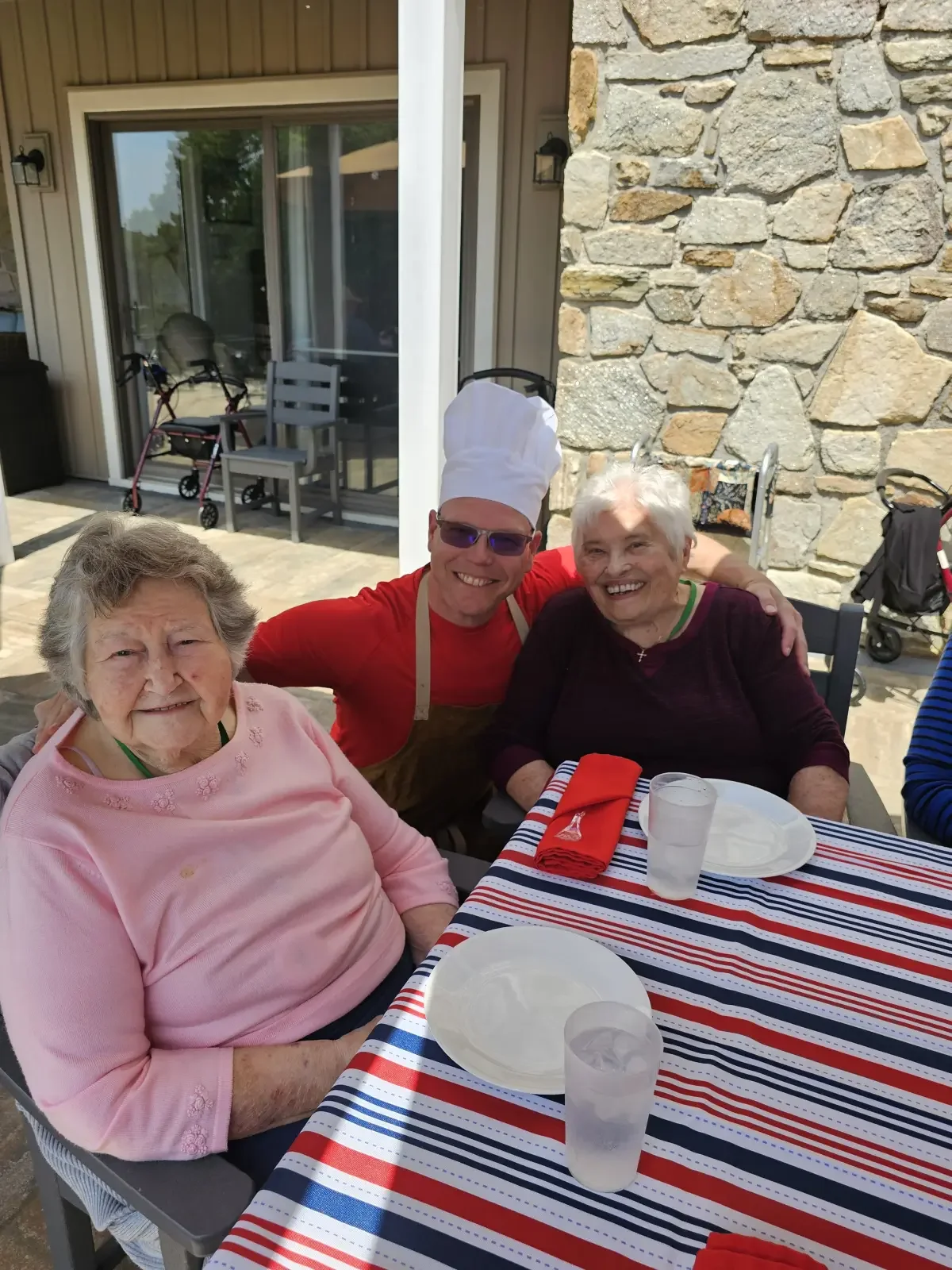 Two smiling residents with the Broad Creek Manor chef, enjoying a meal together at a cozy table on the patio