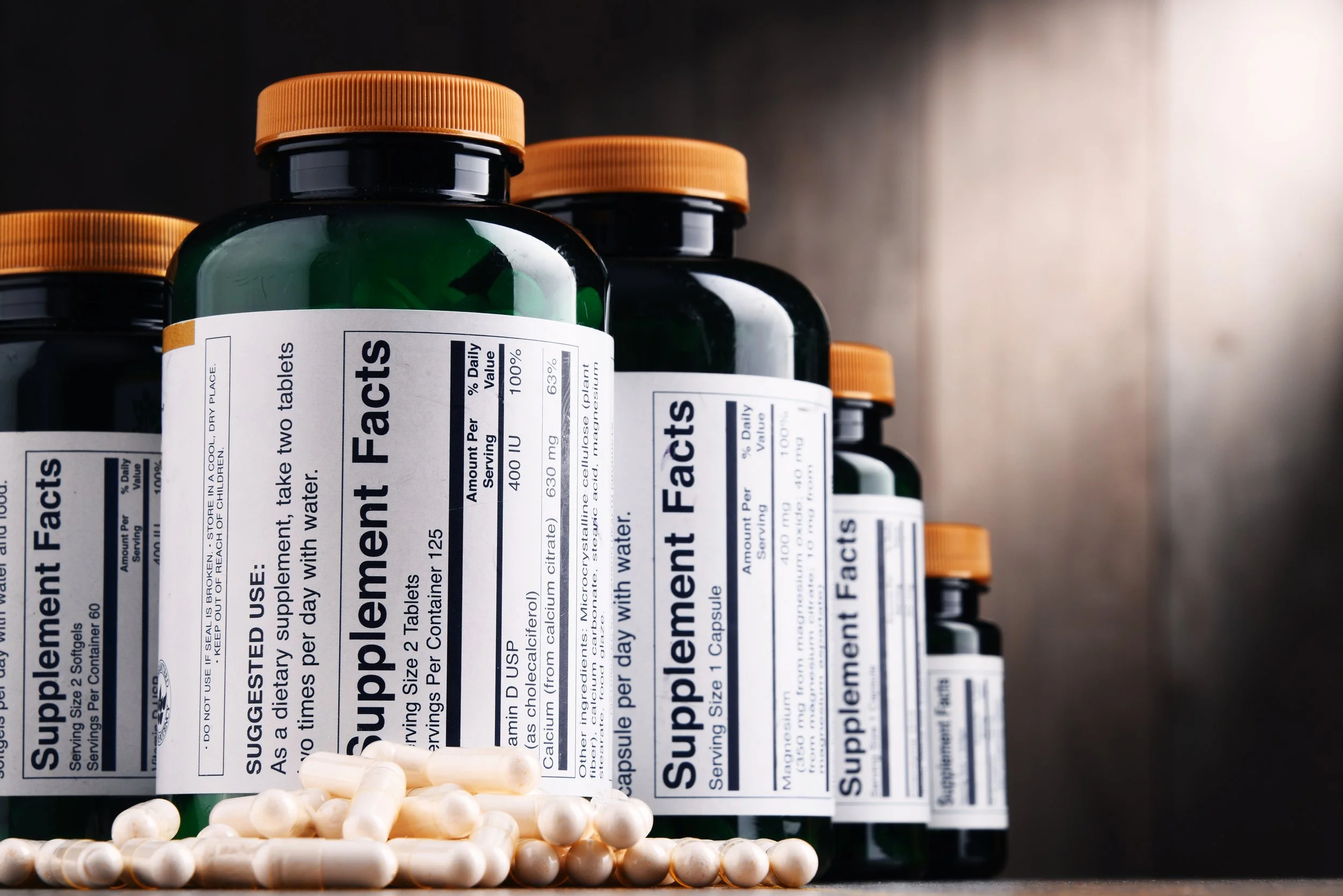 Bottles of dietary supplement tablets with labels titled 'Supplement Facts' and a pile of white capsules in front.