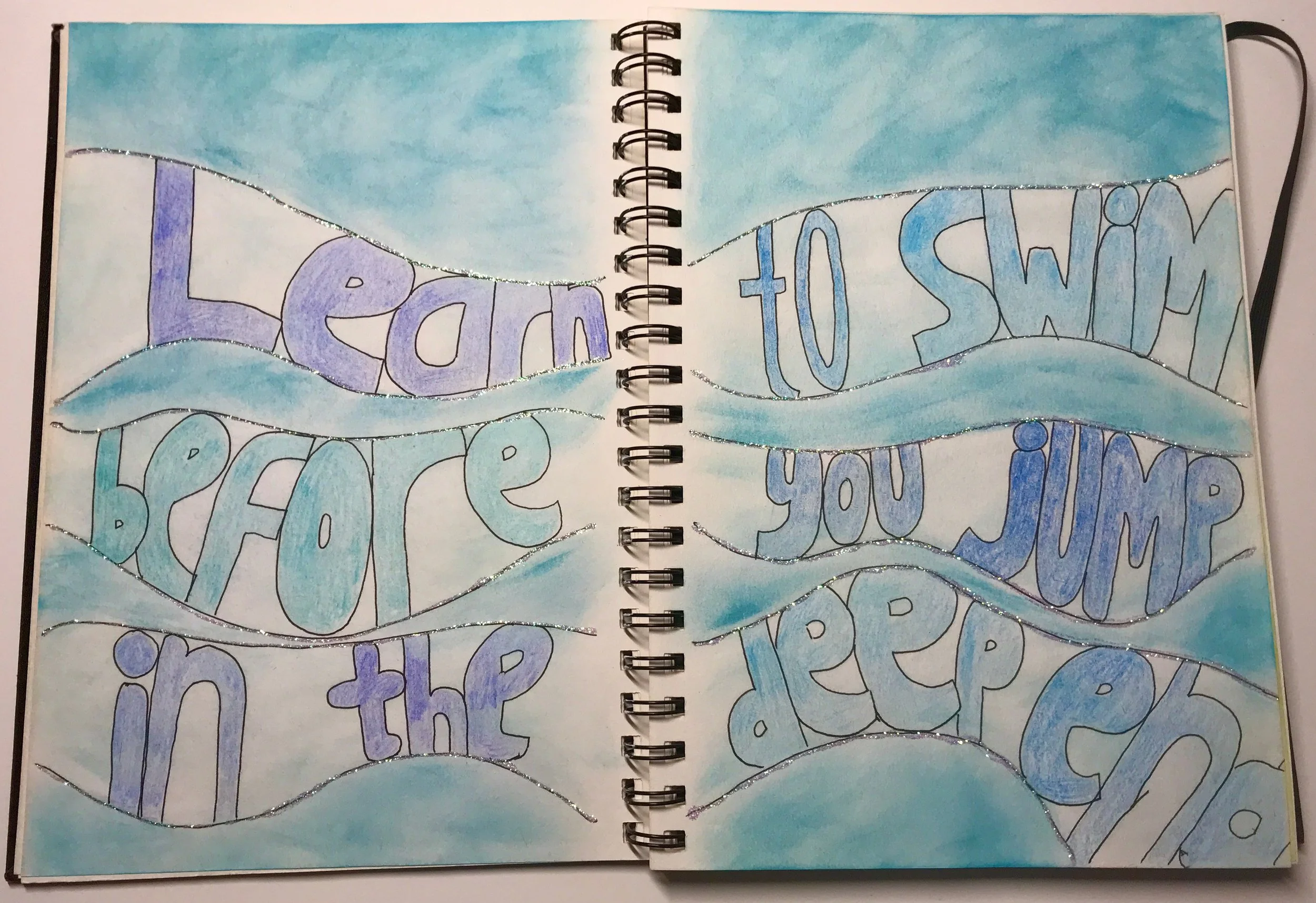 An open ringbound journal, with the words 'Learn to swim before you jump in the deep end' written in wave shapes across the page. Various colours of blue used.