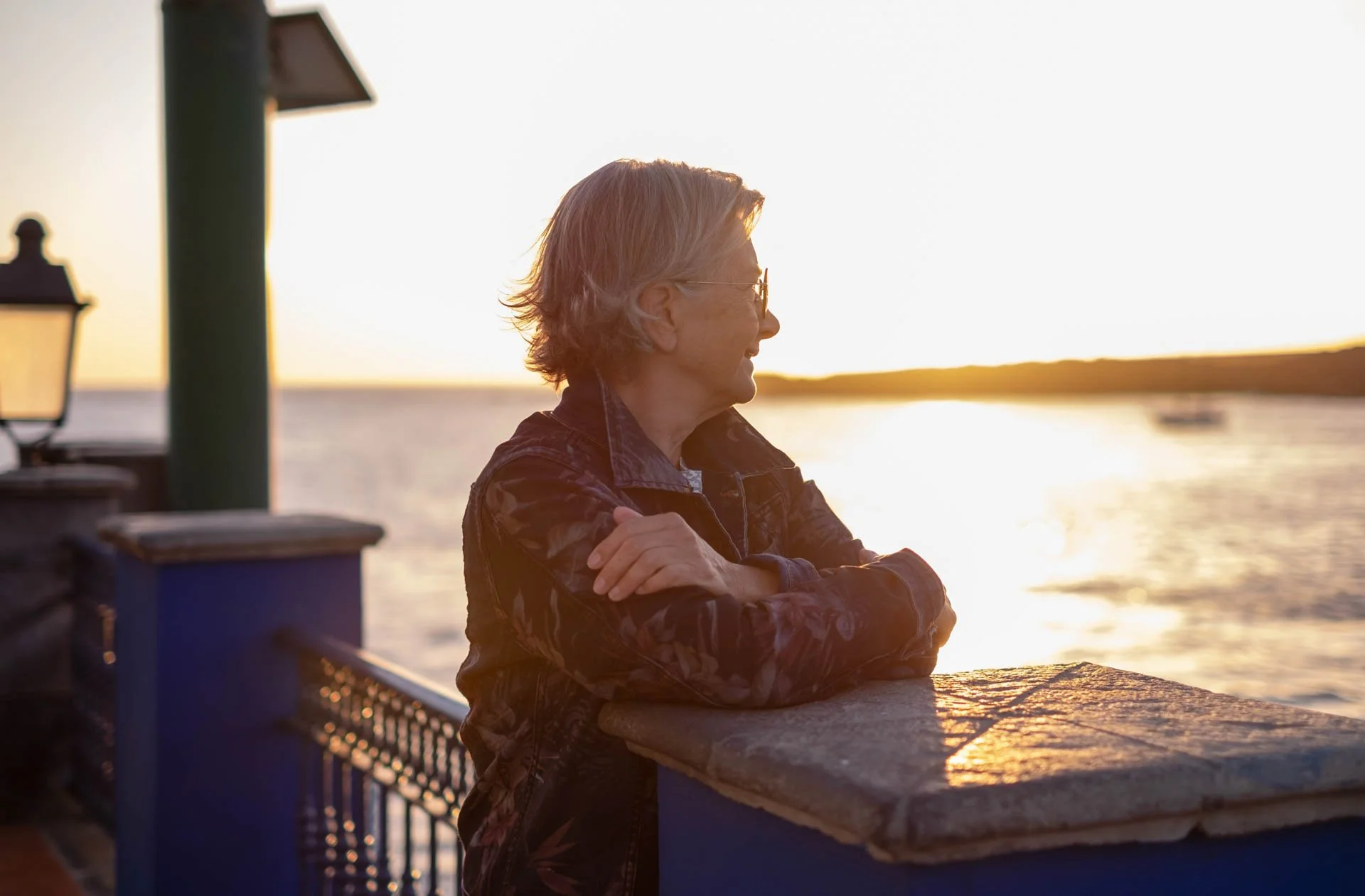 A woman with gray hair and glasses stands by a railing at a waterfront during sunset, looking out at the water.
