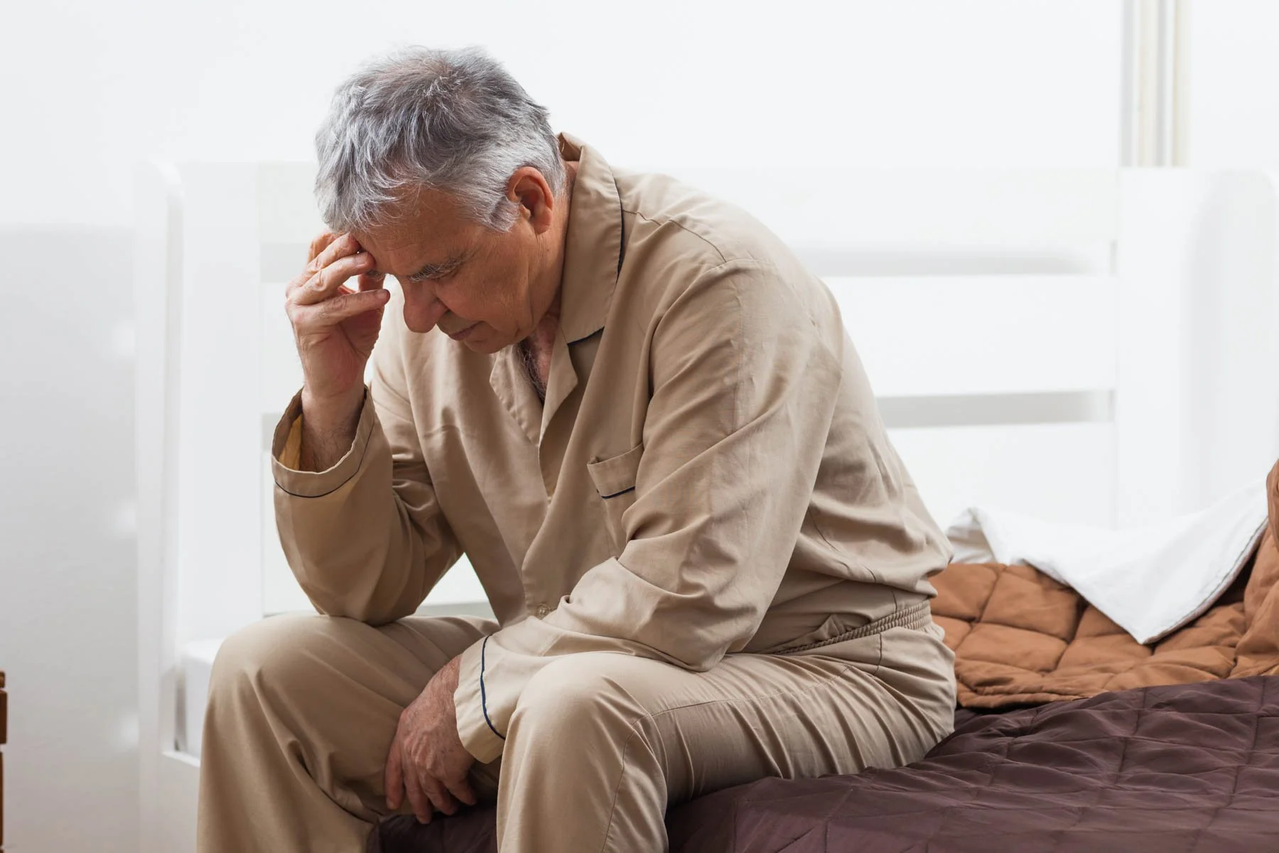 An elderly man with gray hair, sitting on a bed in pajamas, holding his head with one hand, appearing distressed or in pain.