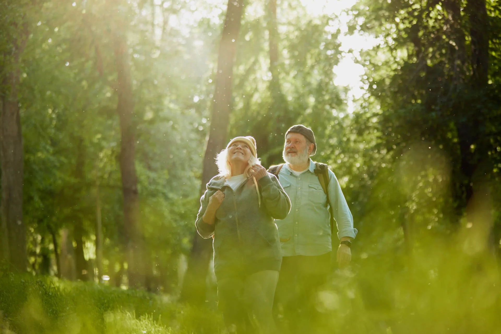 An older couple hiking through a sunlit forest, enjoying nature.