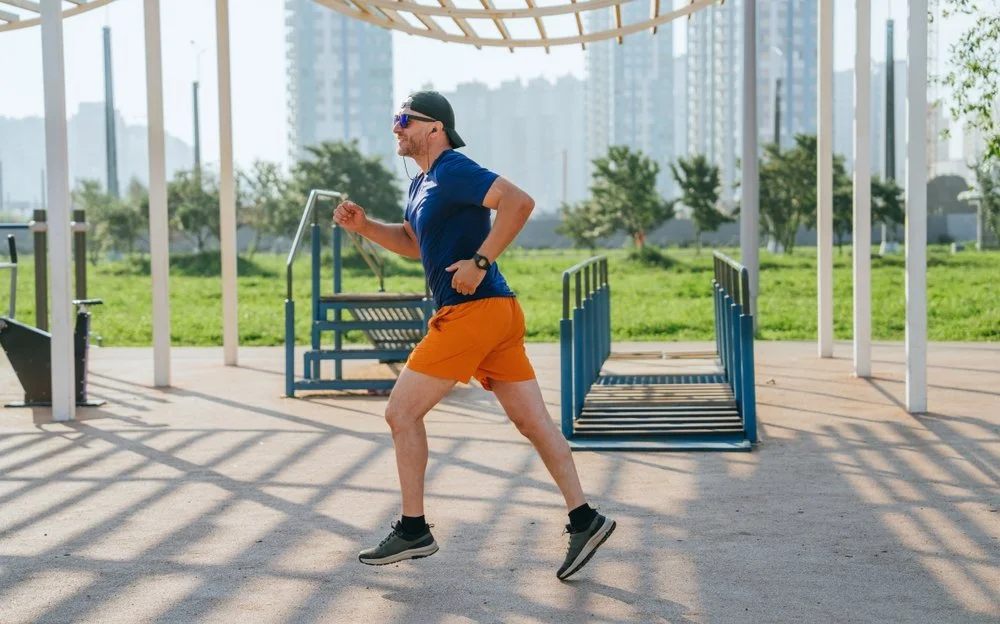 Man running outdoors at a park on a sunny day, wearing athletic clothes, sunglasses, and a black cap.