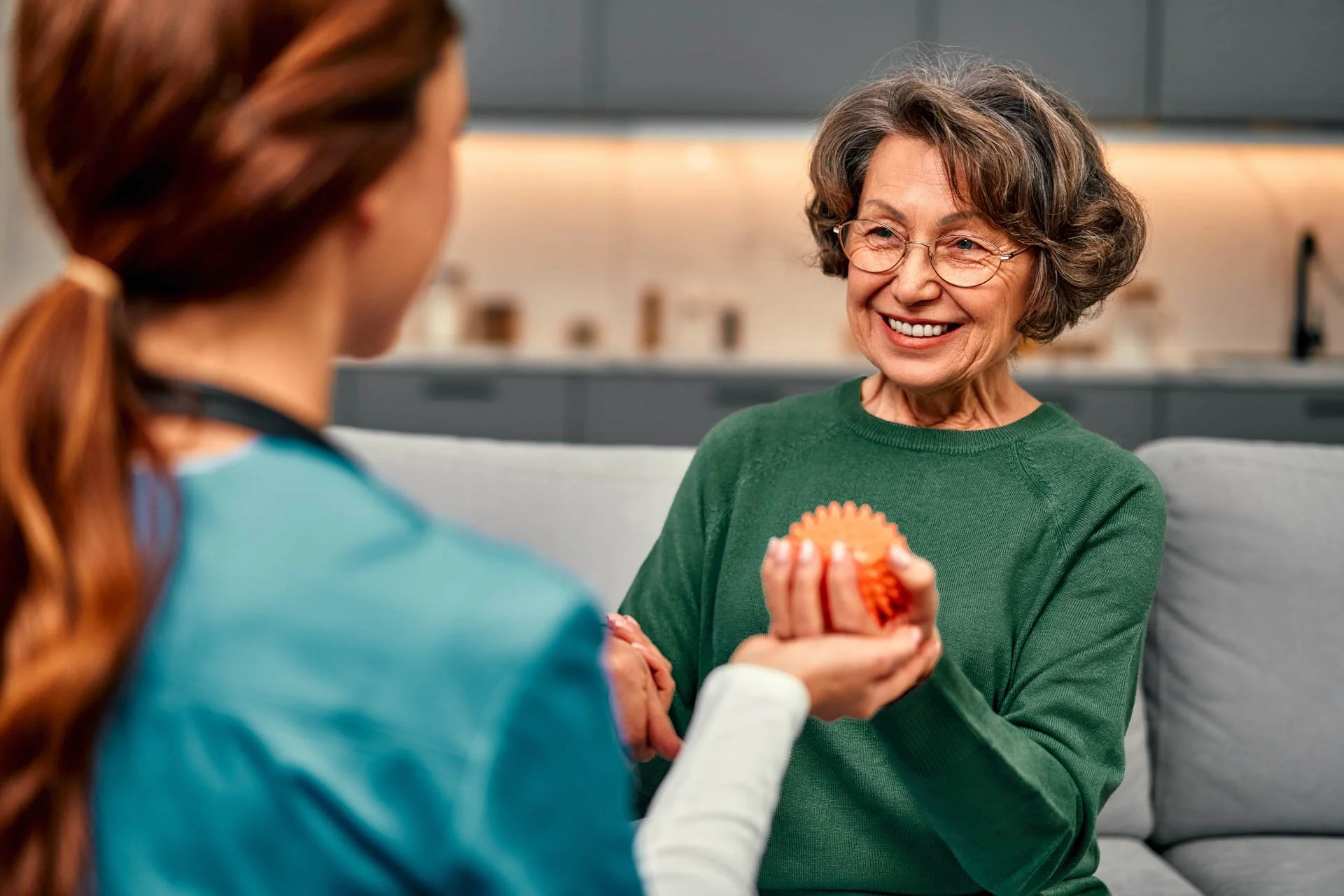 Elderly woman in green sweater smiling and handing a silicone cupcake mold to a female healthcare worker in scrubs at a medical or care facility.