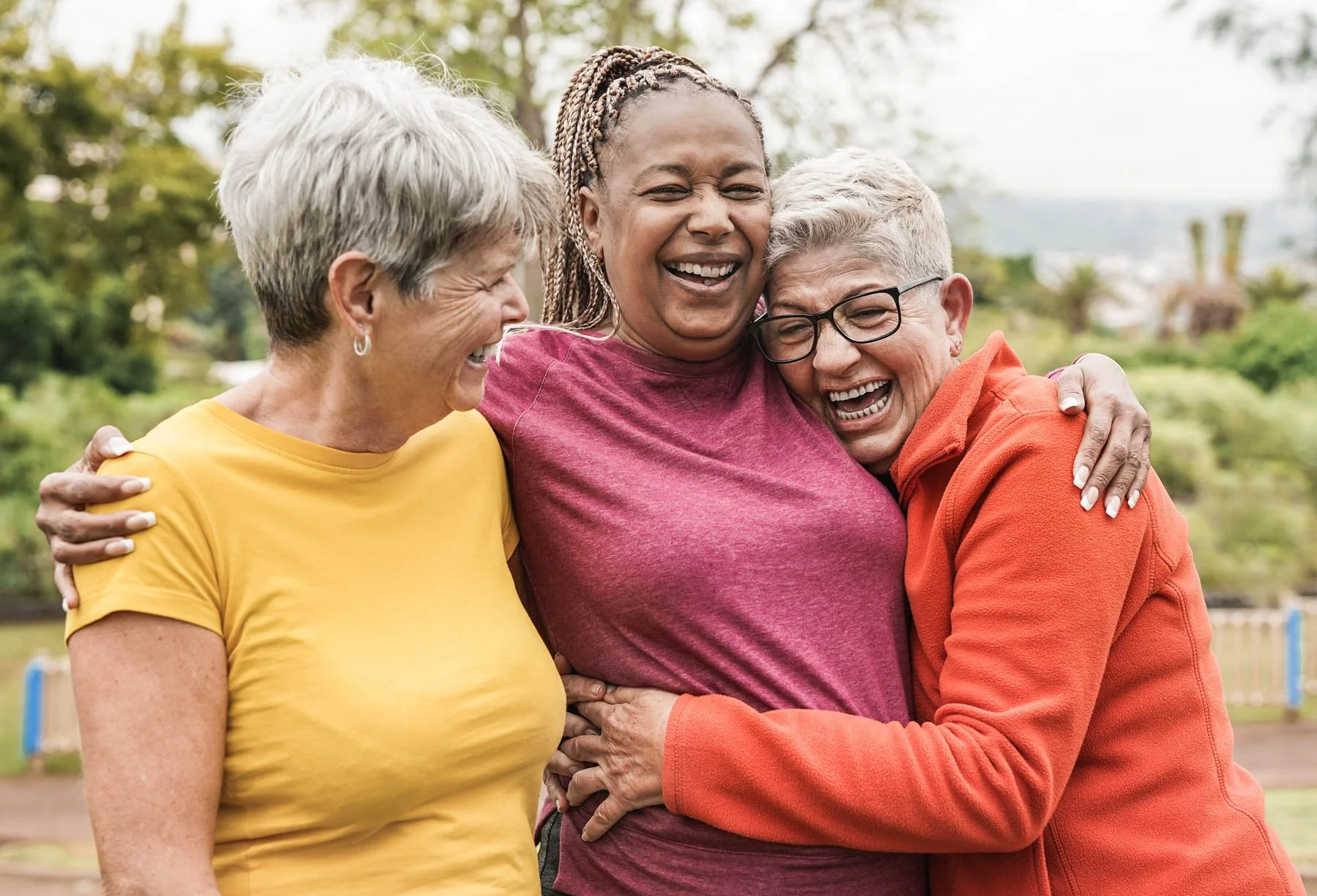 Three women joyfully hugging and laughing outdoors in a park.