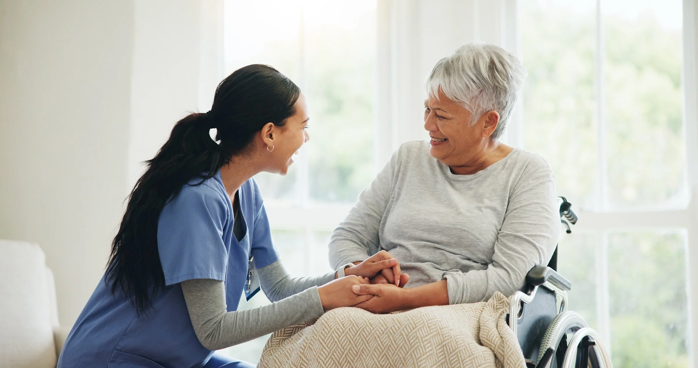 Nursing assistant holding hands of an elderly woman in a wheelchair, both smiling warmly in a bright room.