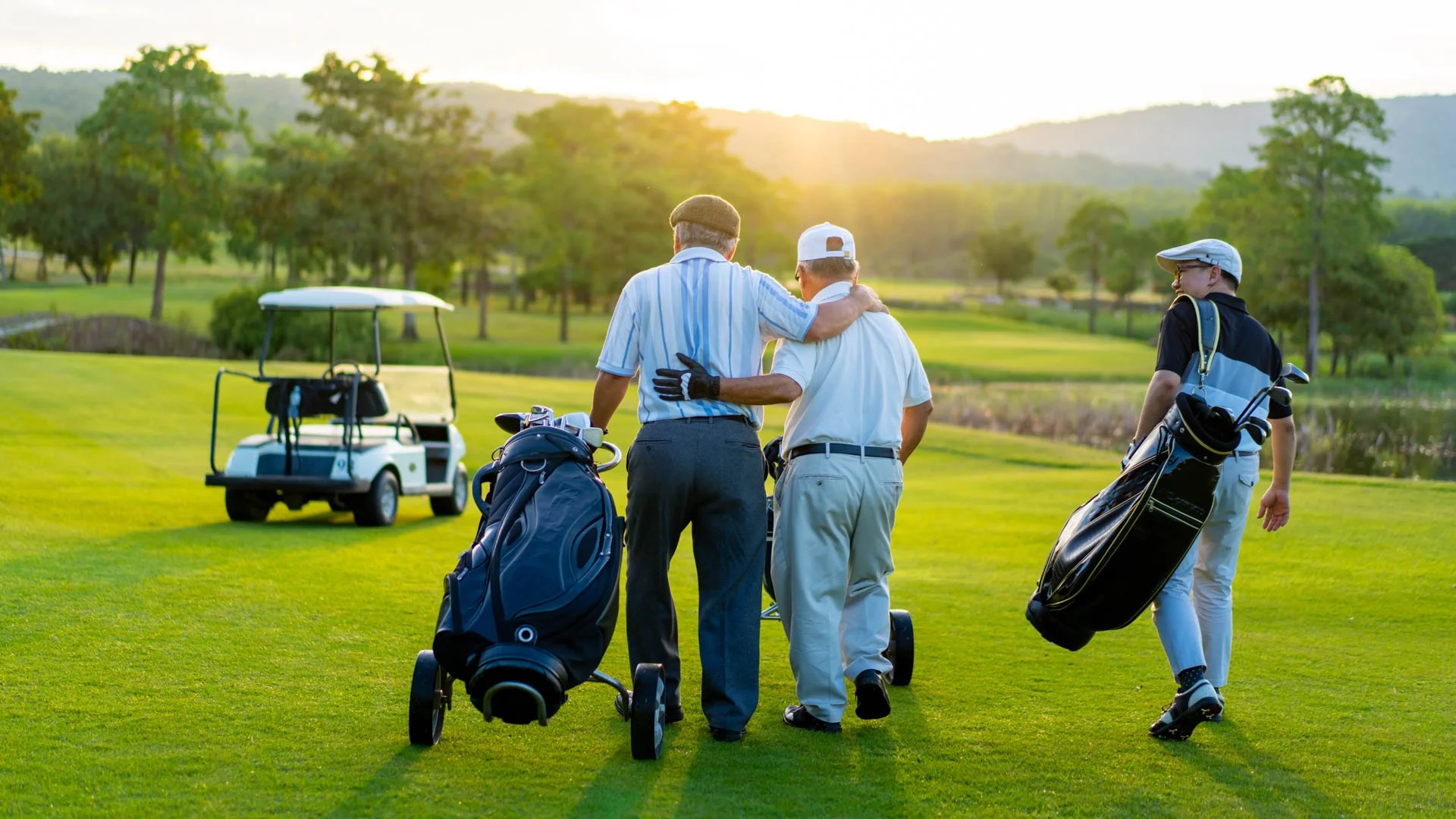 Three men walking on a golf course, with a golf cart and golf bags, during sunset.