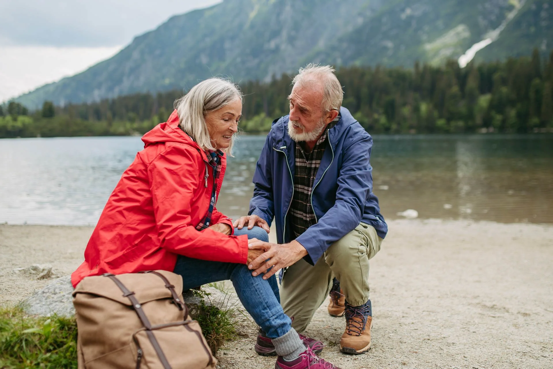 An elderly man and woman sitting on a shore by a lake in a mountainous area, with the man checking the woman's knee, suggesting an incident during outdoor activity.