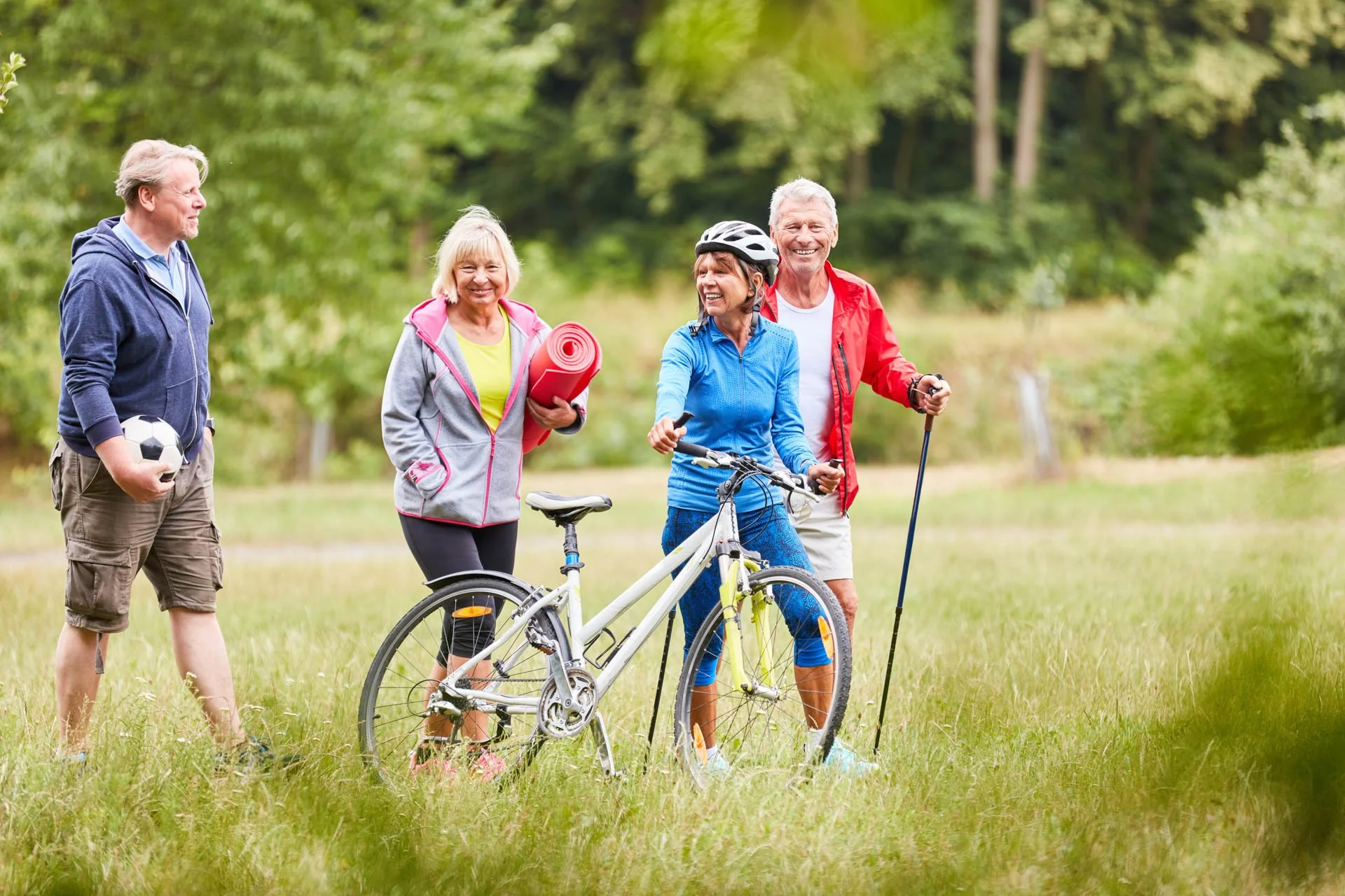Group of five seniors outdoors in a grassy park, smiling and chatting. One woman is wearing a blue jacket with a bicycle, another woman holds a yoga mat, a man holds a soccer ball, and a man in a red jacket holds a walking stick.