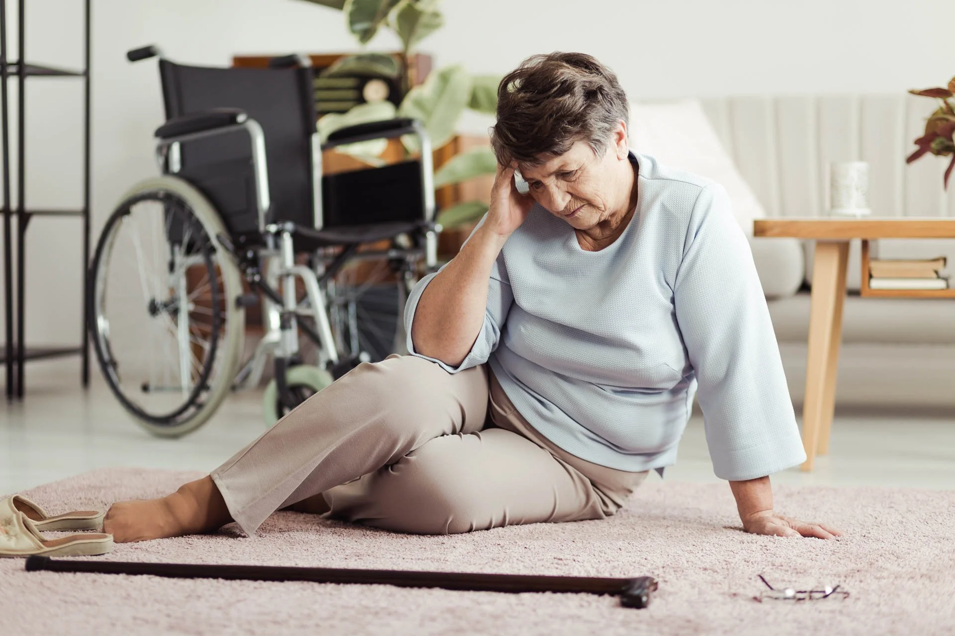 An elderly woman sitting on a carpet, holding her head with one hand, appears distressed. She is barefoot and has a walking cane beside her. In the background, there is a wheelchair and a living room setting with a sofa.