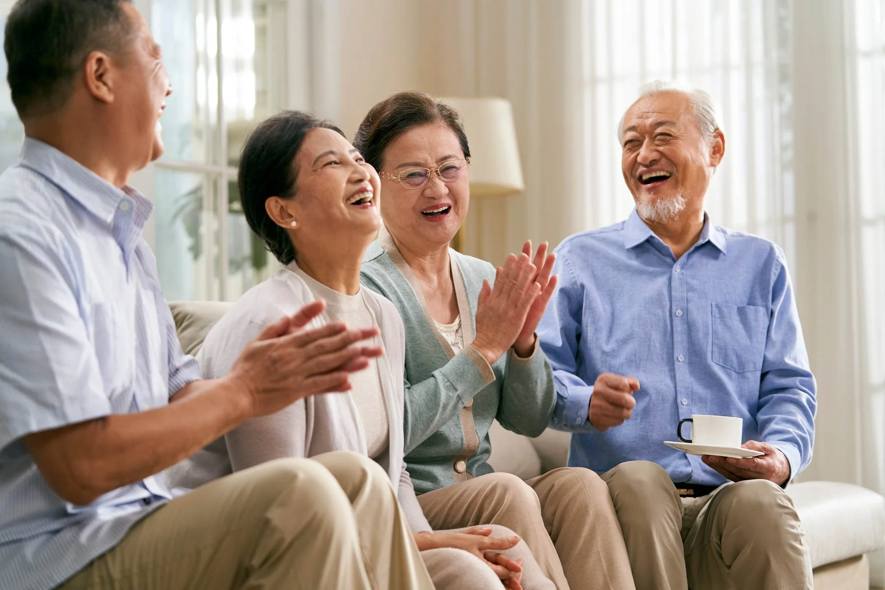 A group of elderly friends and a middle-aged man laughing and clapping together in a living room.