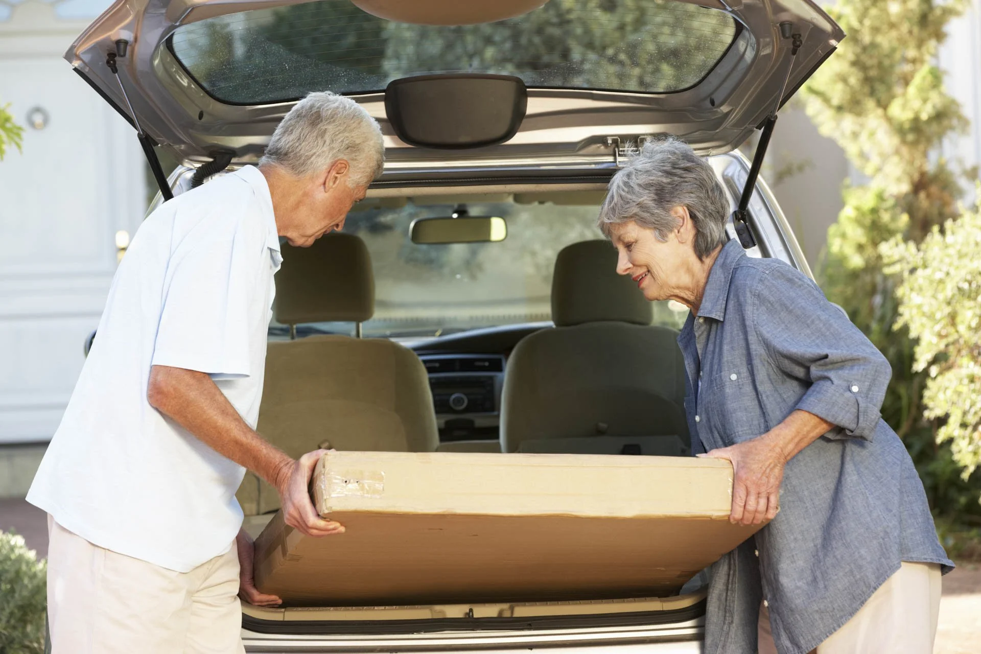 An elderly man and woman loading a large cardboard box into the back of a car.