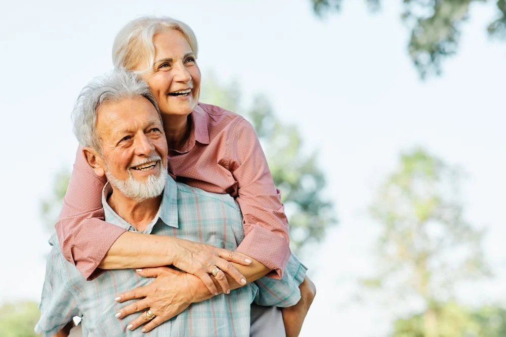 An elderly couple smiling outdoors, the woman on the man's back with her arms around his shoulders.