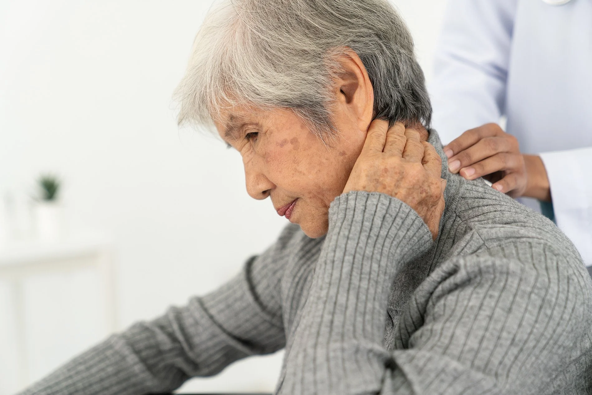 An elderly woman experiencing neck pain while a healthcare professional provides support.