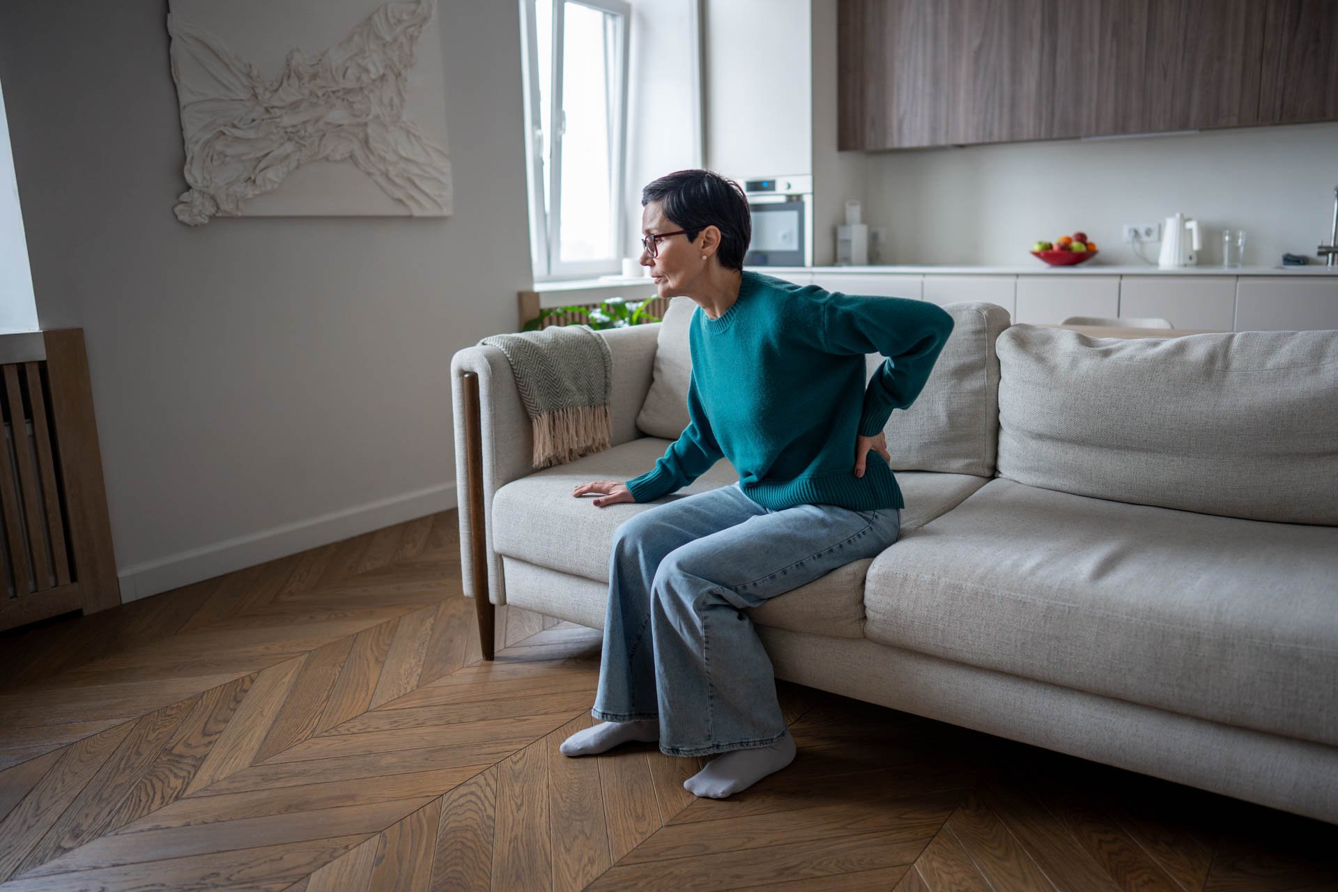 A woman with short dark hair, glasses, wearing a teal sweater and light blue jeans, sitting on a beige sofa with her back pain, supporting her lower back with her right hand in a modern living room.