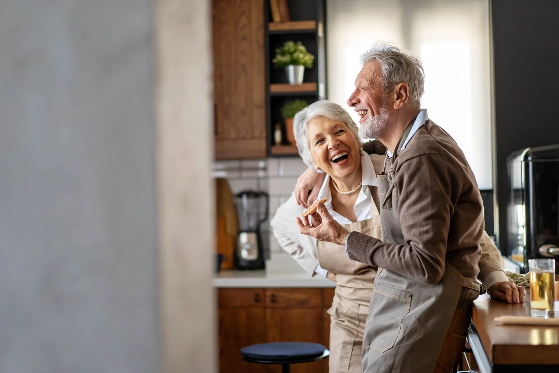 An elderly couple laughing and enjoying themselves in the kitchen, possibly cooking or sharing a moment together.
