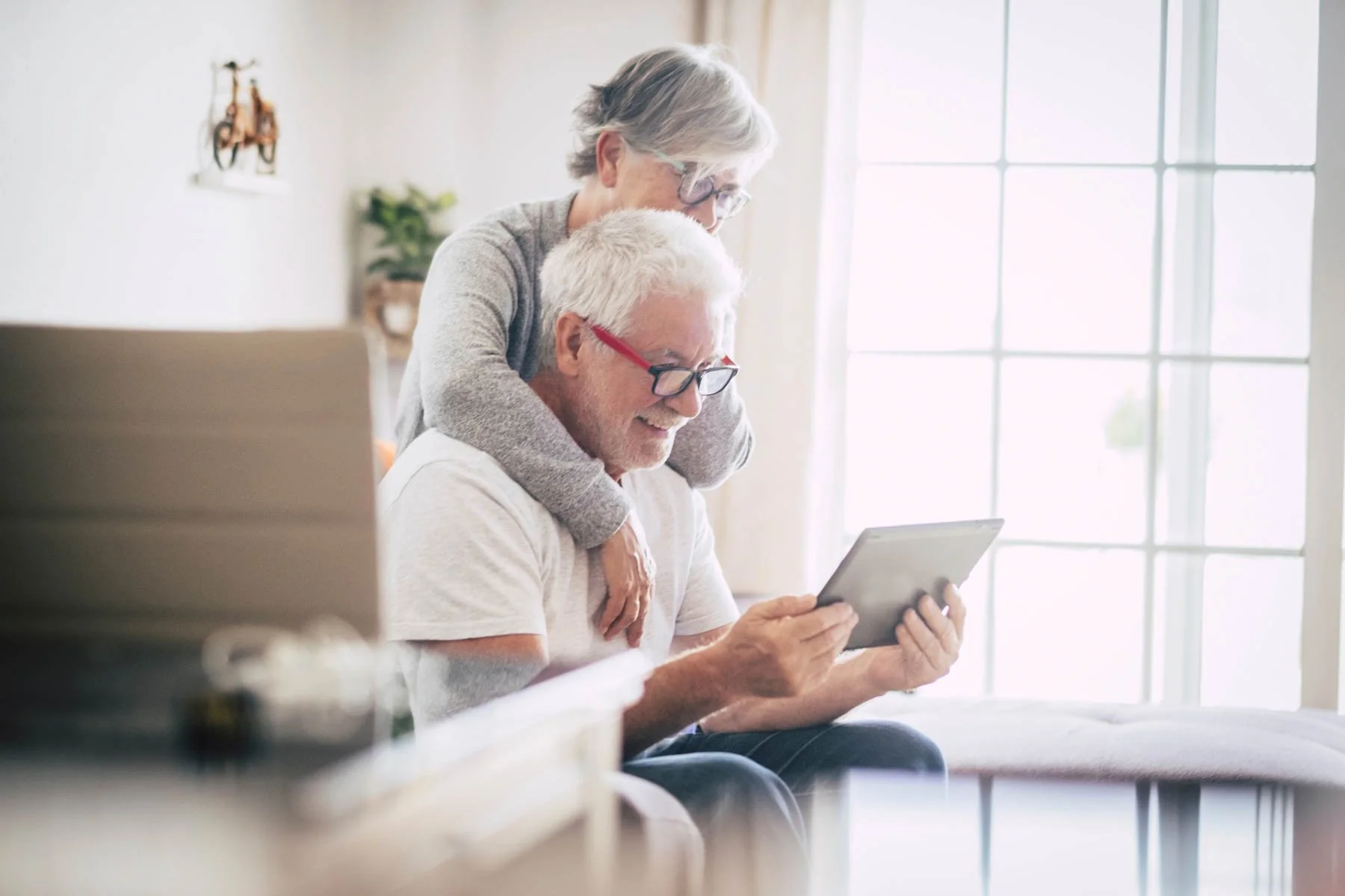 An elderly couple sitting on a sofa, with the woman embracing the man from behind, looking at a tablet together in a bright living room.