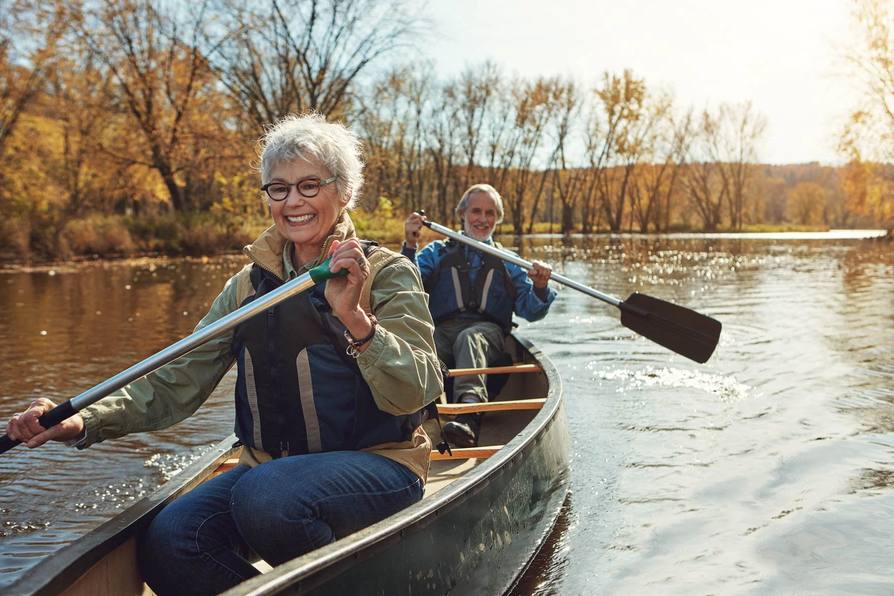 Two older adults, a woman and a man, are paddling a canoe on a river during autumn with trees showing fall colors in the background. They are smiling and seem to be enjoying their outdoor activity.