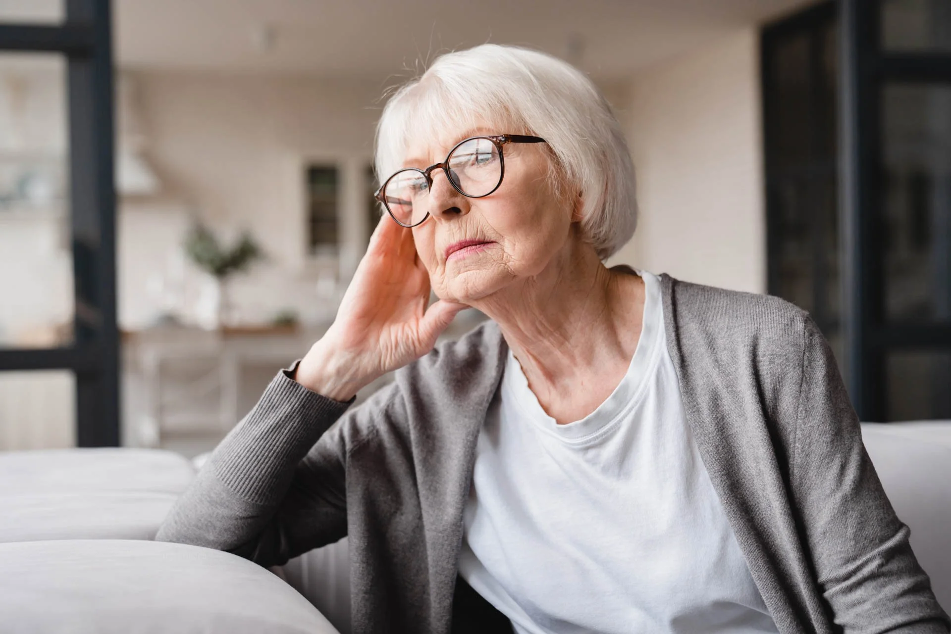 An elderly woman with white hair and glasses, resting her head on her hand while sitting on a sofa in a modern living room.