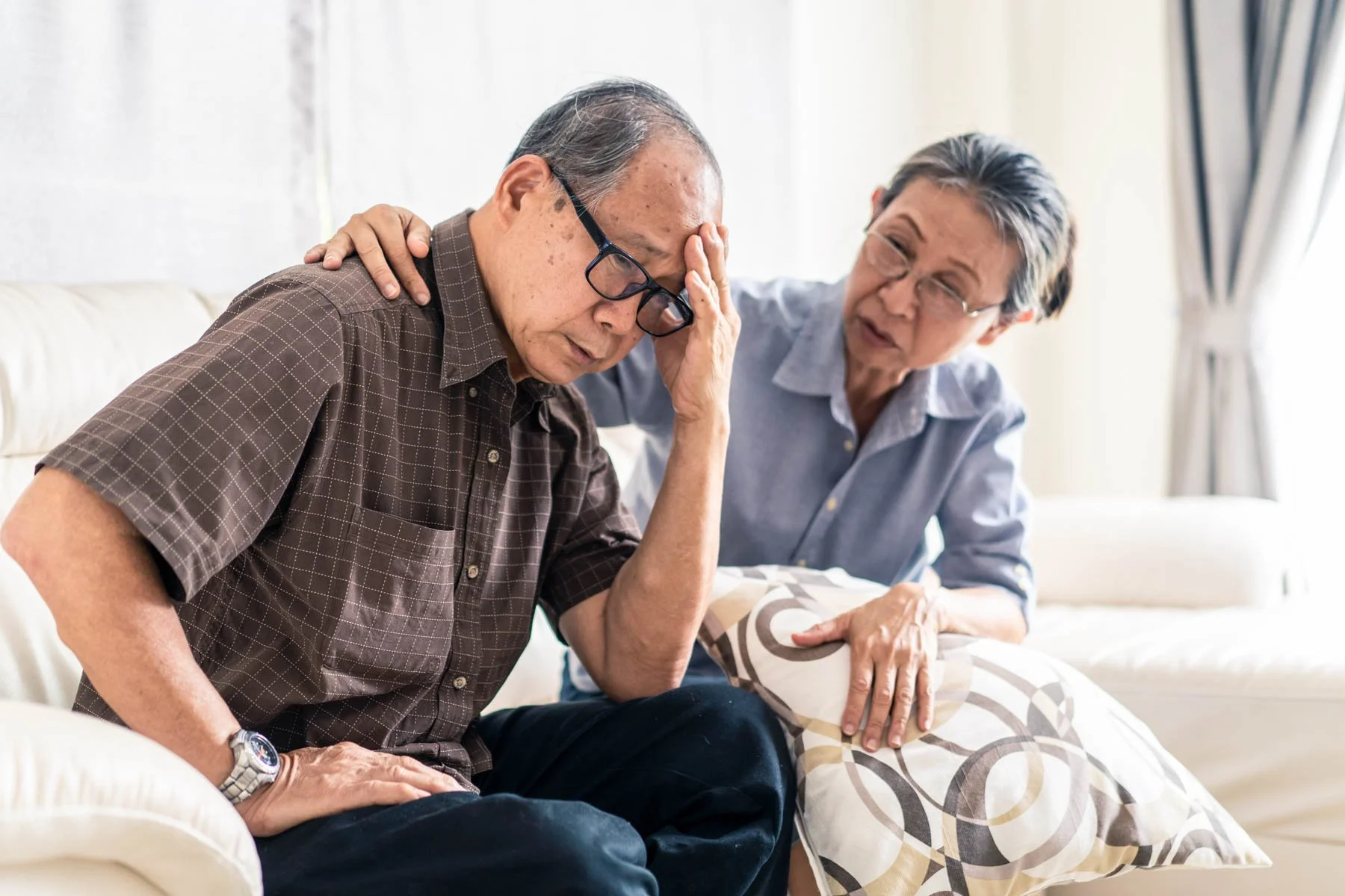 An elderly man with glasses sitting on a sofa holding his head in distress while a woman, possibly his caregiver or family member, places a comforting hand on his shoulder and speaks to him.