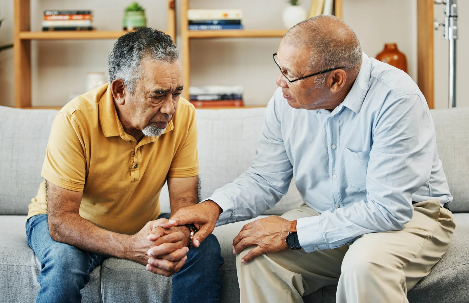 An elderly man wearing a yellow polo shirt sitting on a couch, holding hands with a middle-aged man in glasses, a white shirt, and khaki pants, in what appears to be a comforting moment in a living room.
