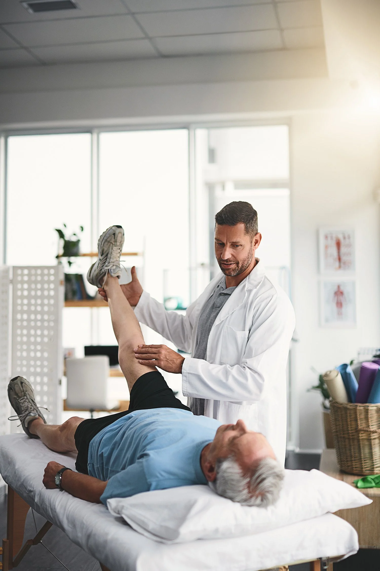 A healthcare professional, wearing a white coat, examines an elderly man lying on a medical examination table, lifting his left leg for assessment in a well-lit clinic or physical therapy office.