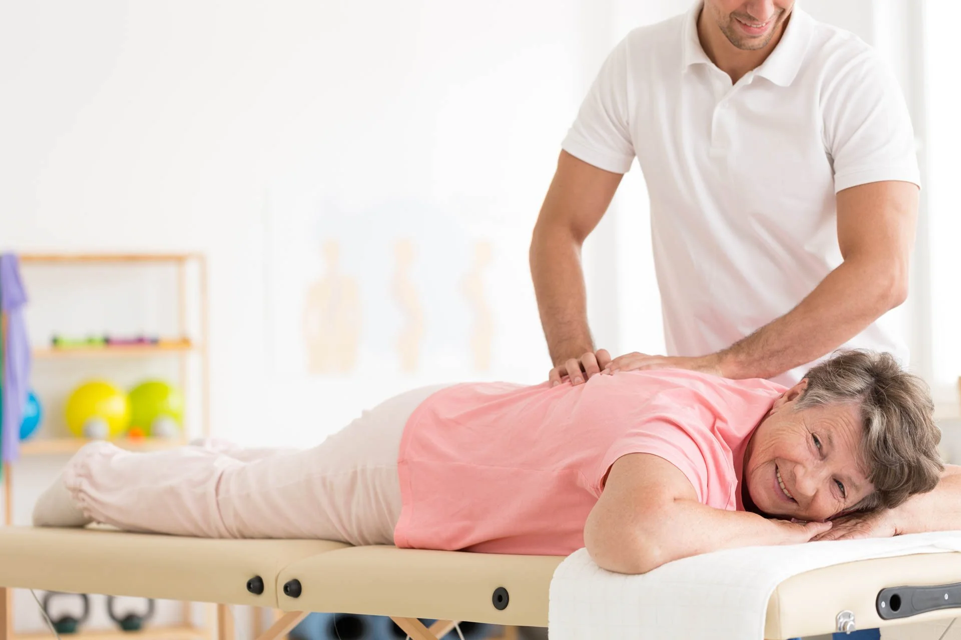 A male physical therapist giving a back massage to an older woman lying face down on a massage table in a wellness or therapy session.