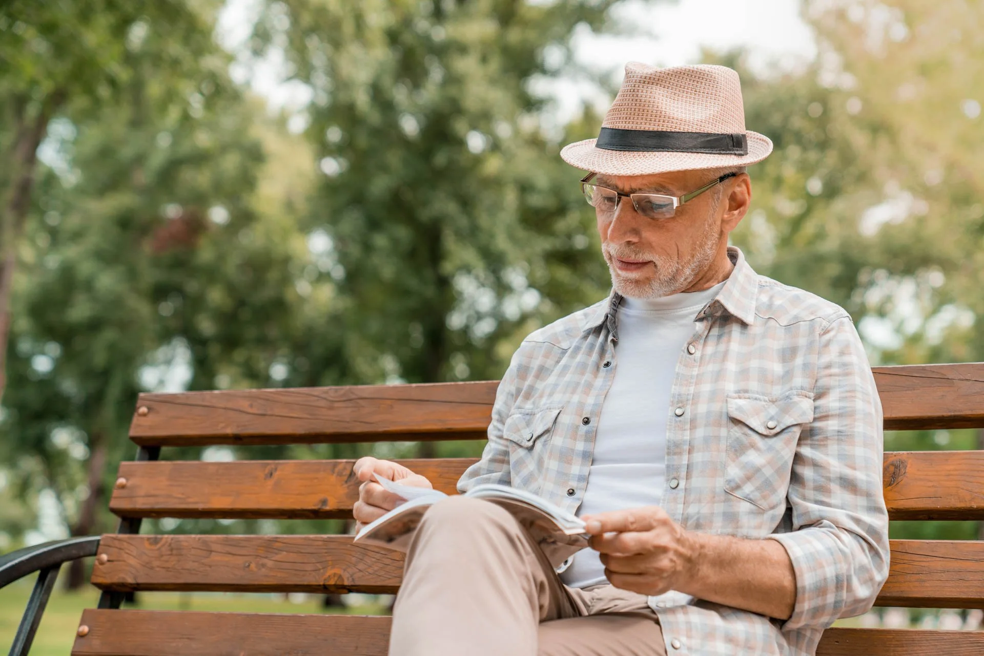 An elderly man with glasses and a hat sitting on a park bench, reading a book surrounded by green trees.