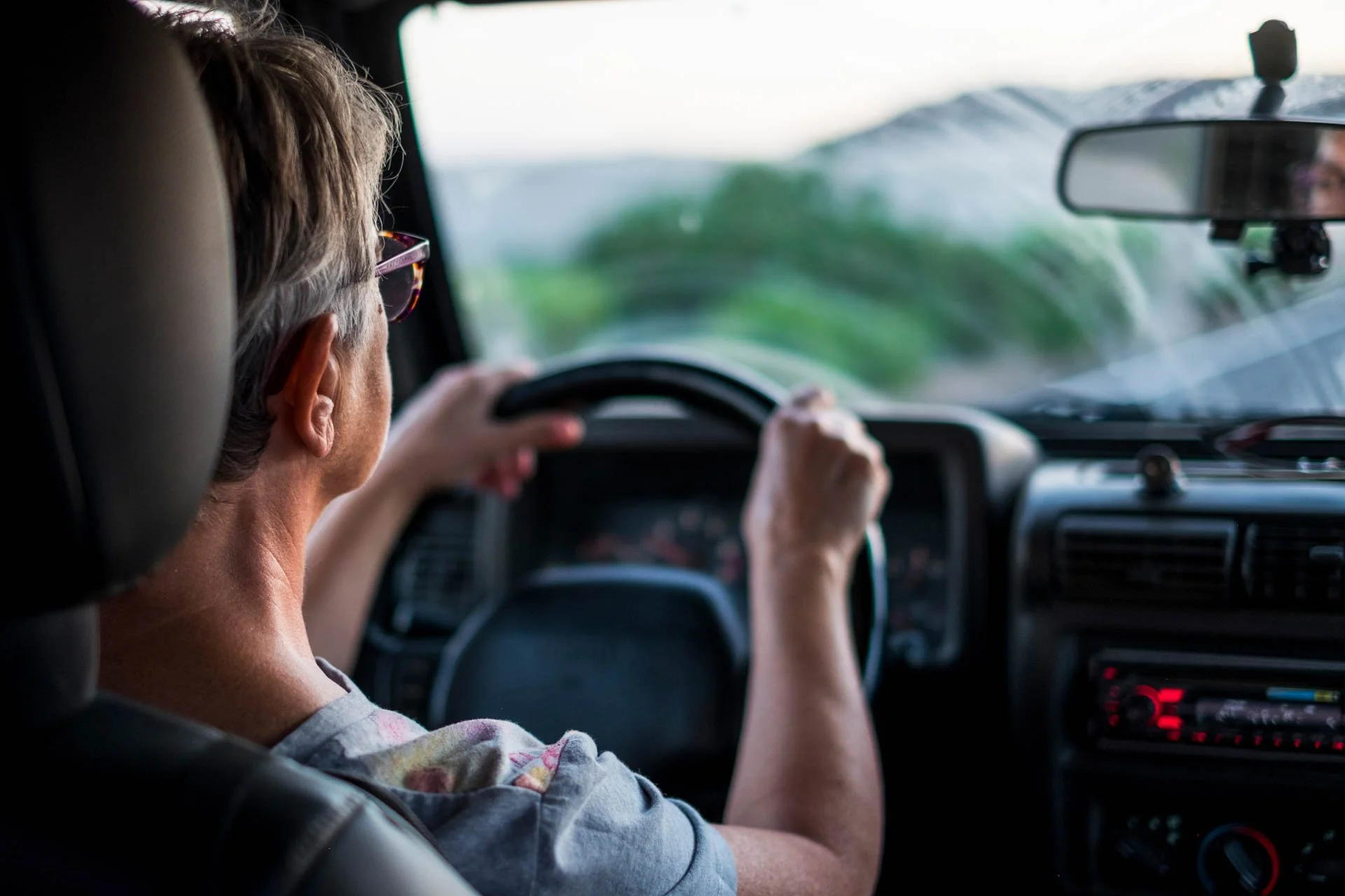 An older woman with short gray hair and glasses driving a vehicle, with a blurred landscape seen through the windshield.