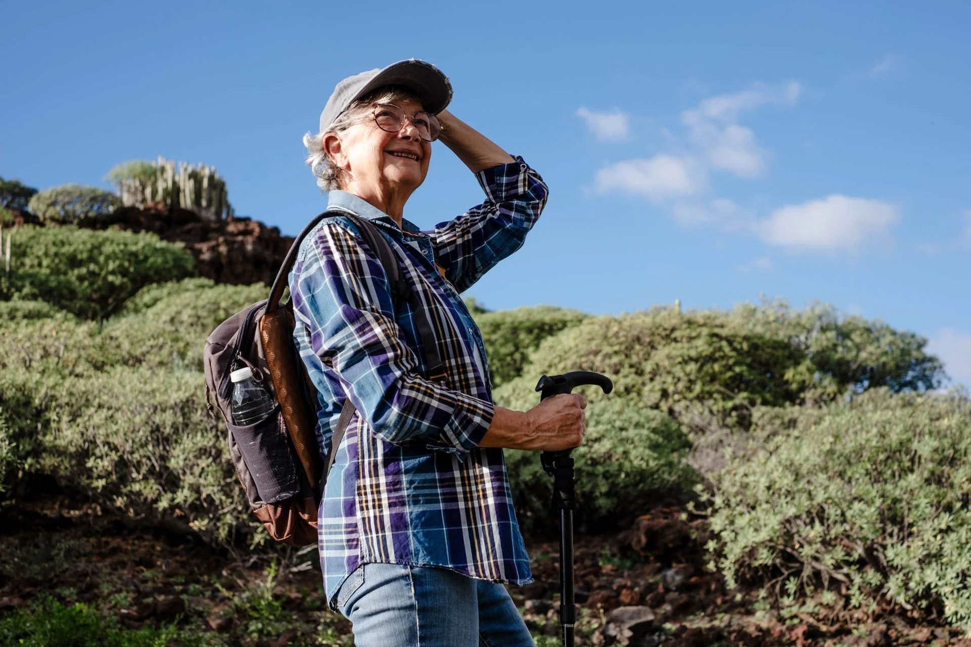 An elderly woman with gray hair, glasses, wearing a hat, plaid shirt, and jeans, stands on a trail with a walking stick outdoors under a blue sky, smiling and holding her hat.