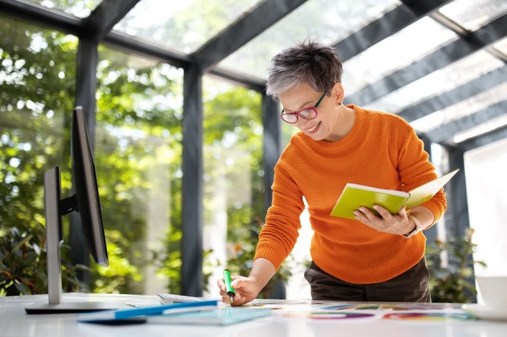 Woman with short gray hair and glasses smiling while standing at a table, holding a yellow notebook and a green marker, in a bright greenhouse or conservatory with large windows and greenery outside.