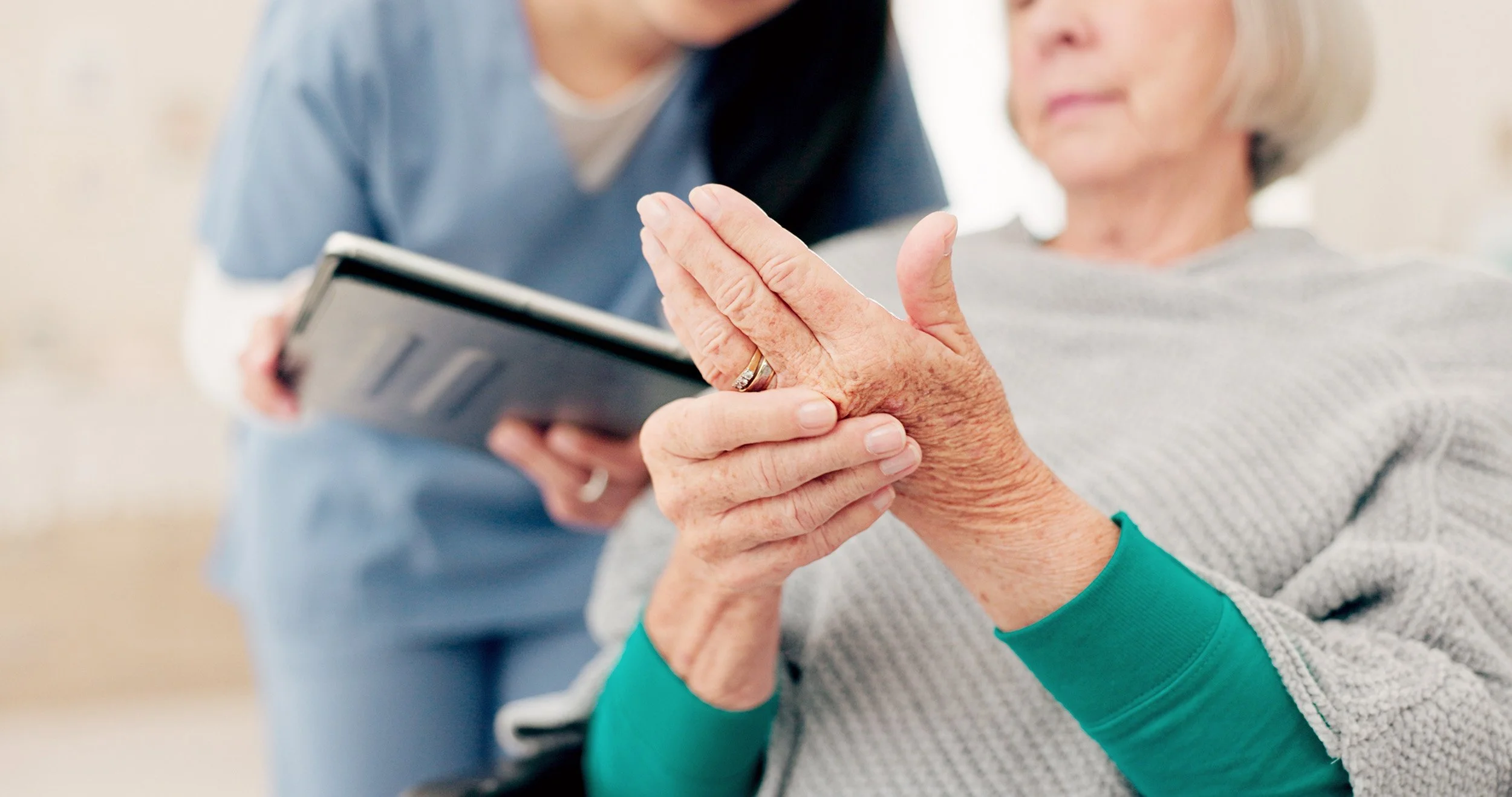 A caregiver assisting an elderly woman with her hands, while holding a tablet.
