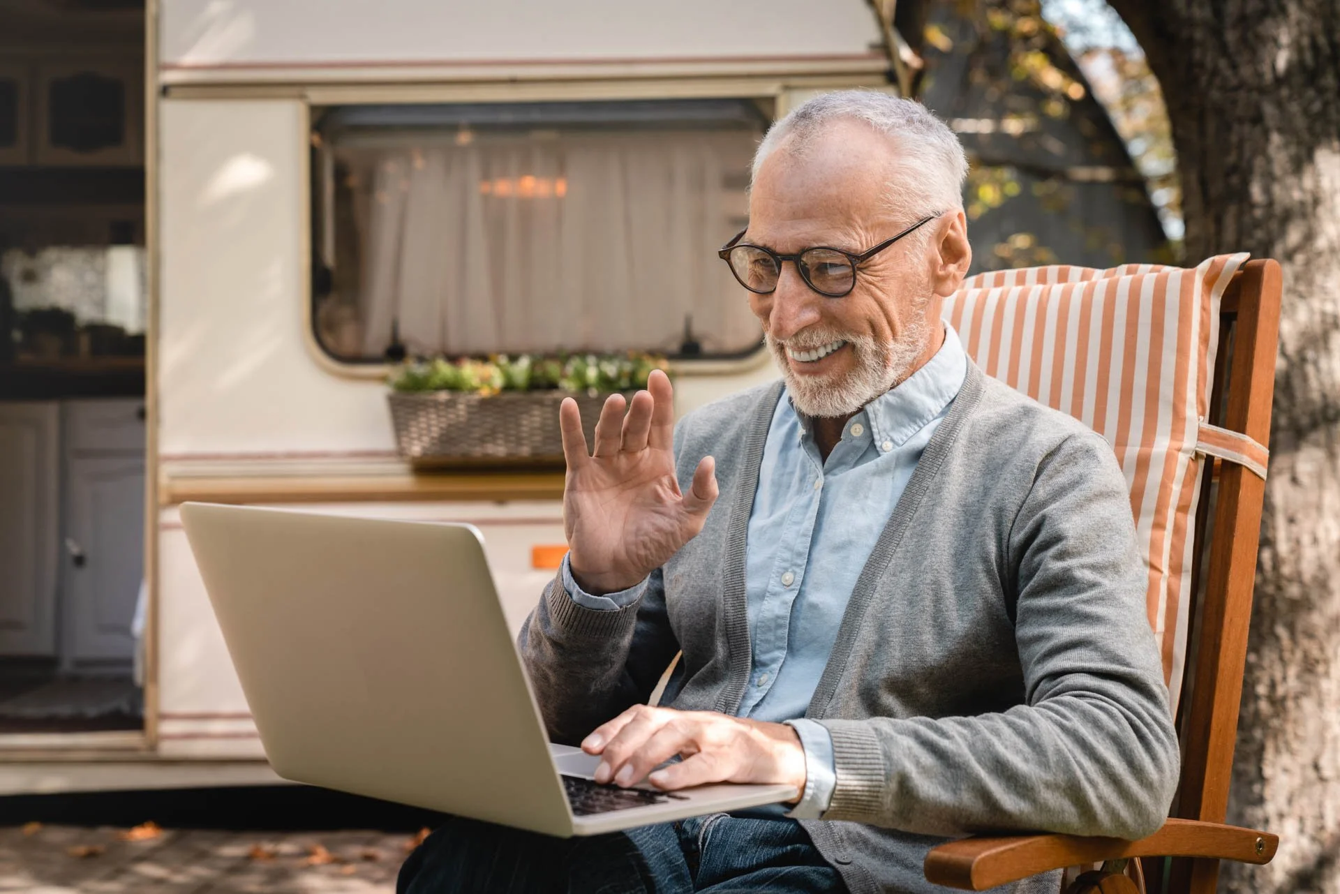 An elderly man with gray hair, beard, and glasses, smiling and waving while sitting in a striped outdoor chair and using a silver laptop, with a vintage camper in the background.
