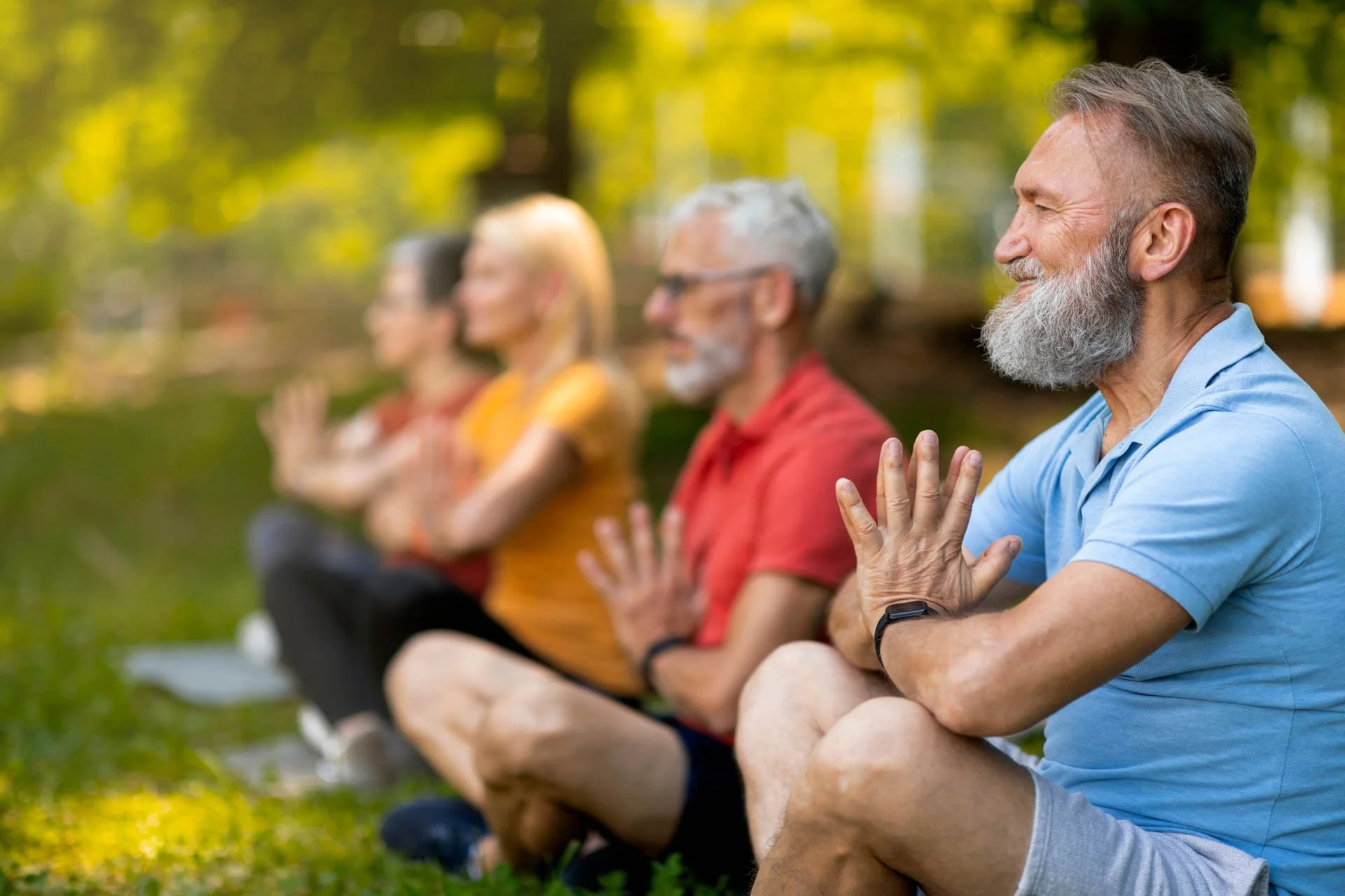 A group of four people practicing yoga outdoors in a park during daytime, sitting cross-legged and meditating with their hands in a prayer position.