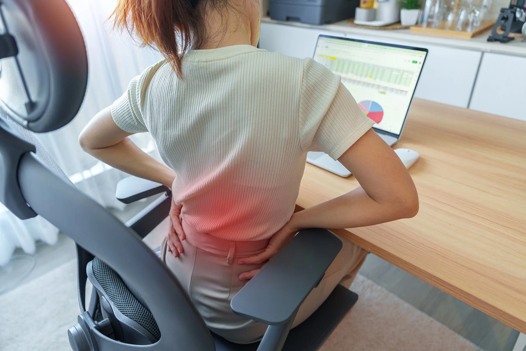 A person sitting in an office chair at a desk, experiencing back pain, with a laptop open displaying graphs and charts.