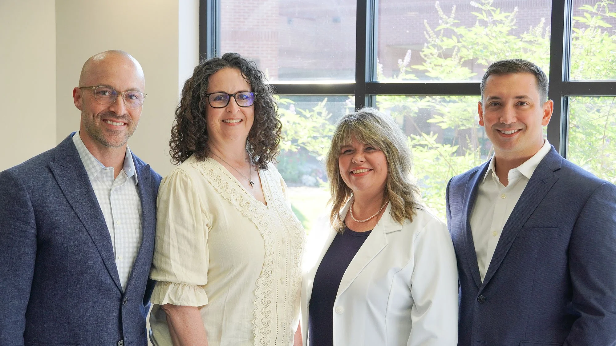 Four professionals standing indoors near large windows with greenery outside, smiling at the camera.