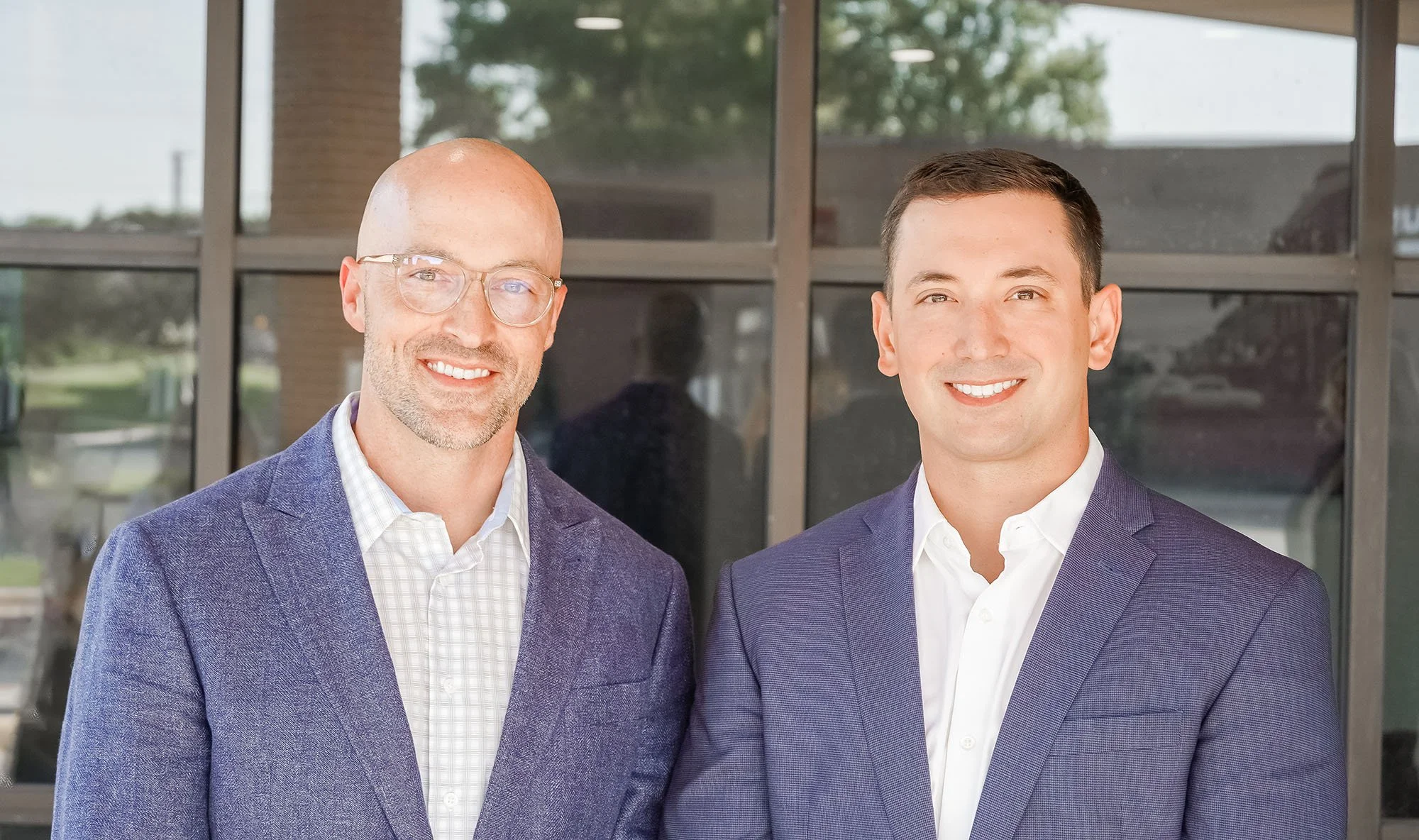 Two men in suits smiling outside a building with large windows.