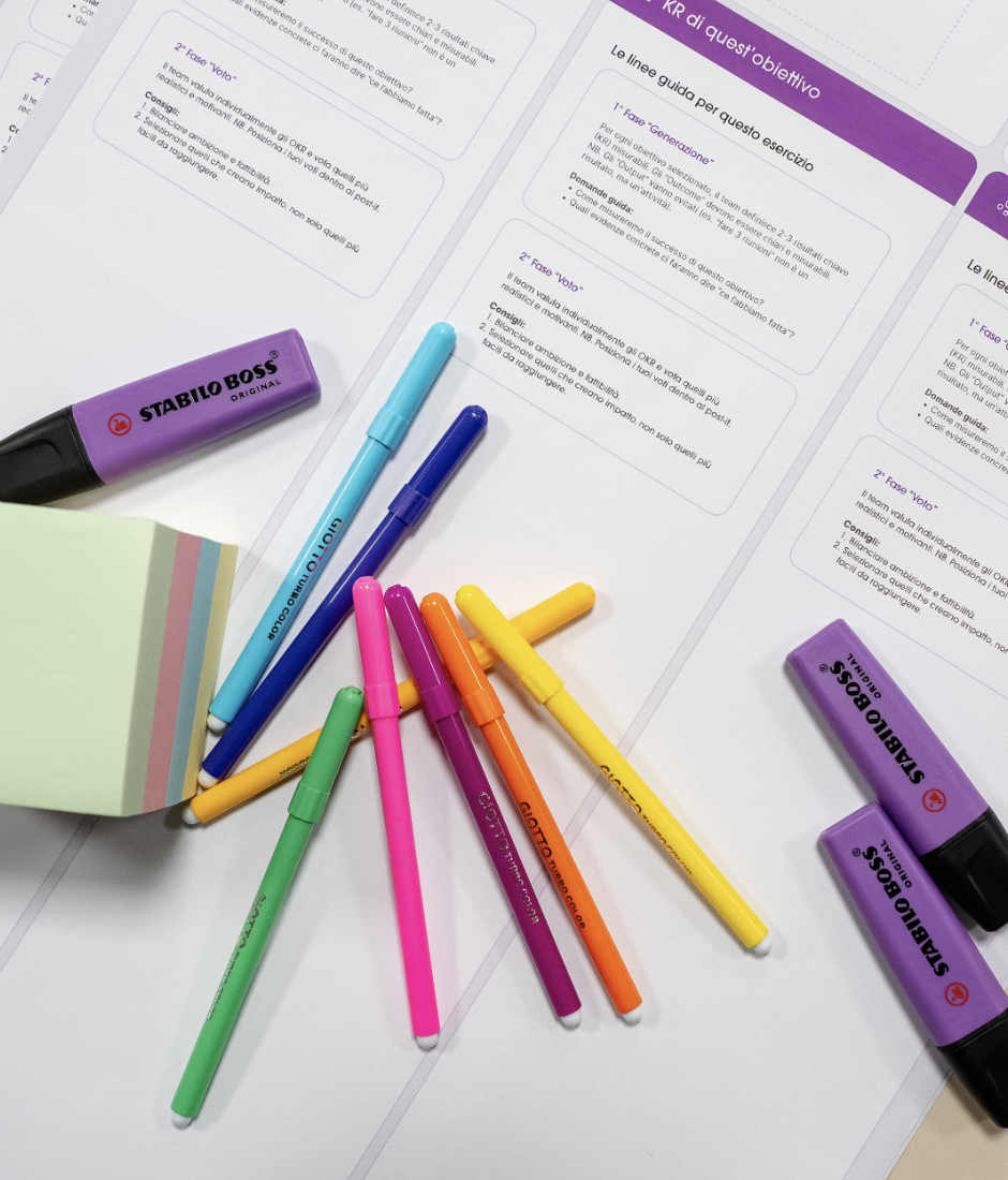 A collection of colorful pens, purple highlighters, sticky notes, and printed documents with text in Italian on a desk.