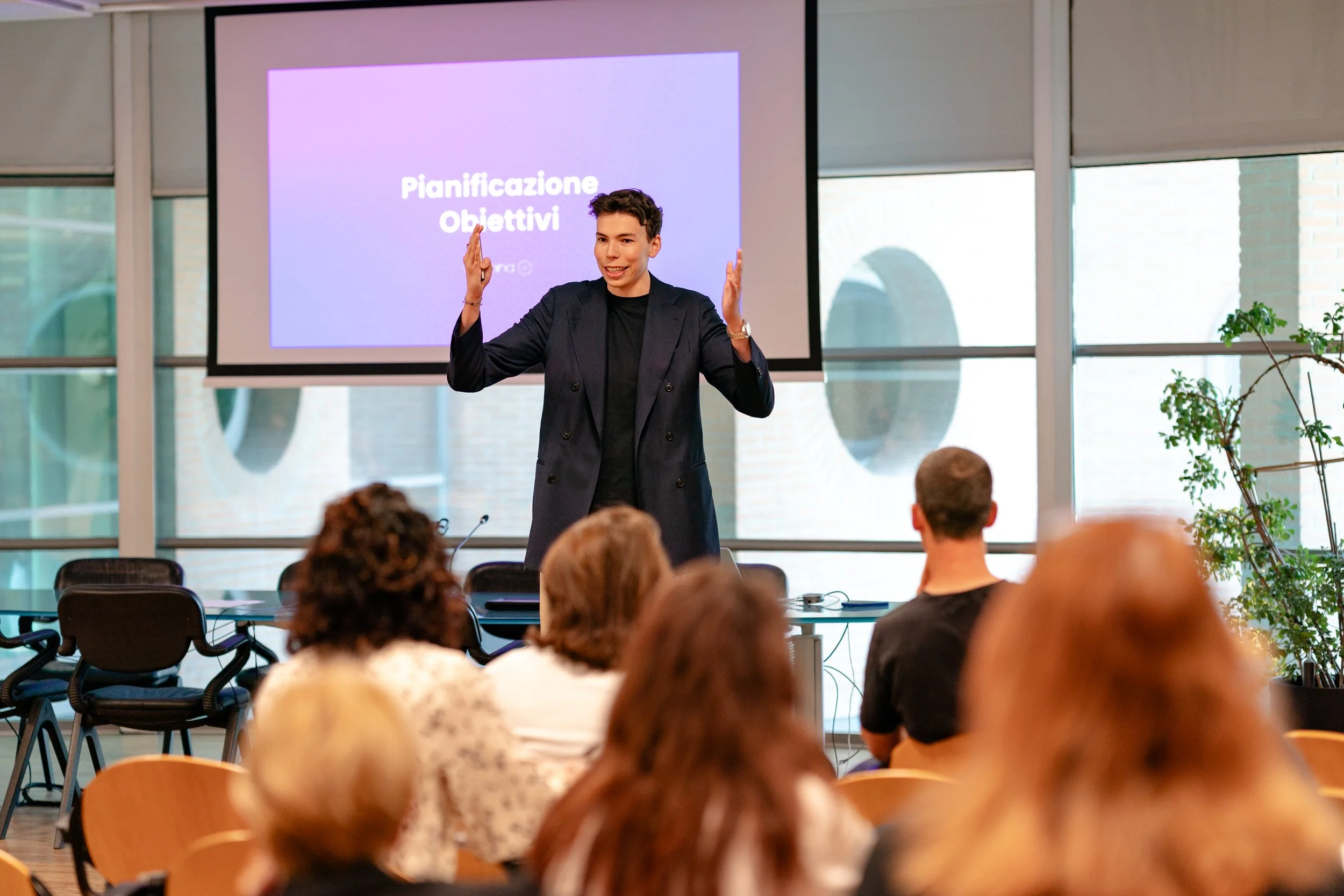 A woman in a dark blazer gives a presentation to an audience in a conference room with large windows, a potted plant, and a screen displaying the words 'Pianificazione Obiettivi'.