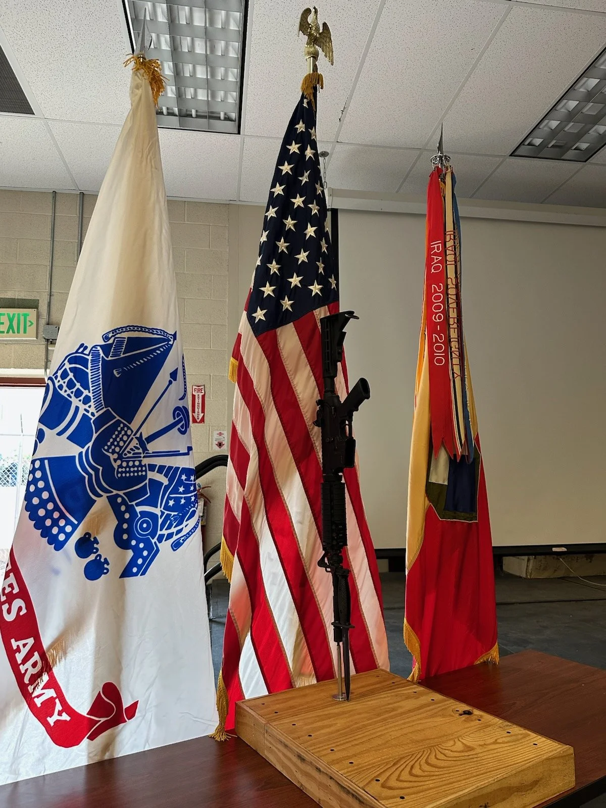 Four flags standing behind a mounted firearm on a wooden stand inside a room, including the United States flag, a firefighting flag, and an American Legion flag.