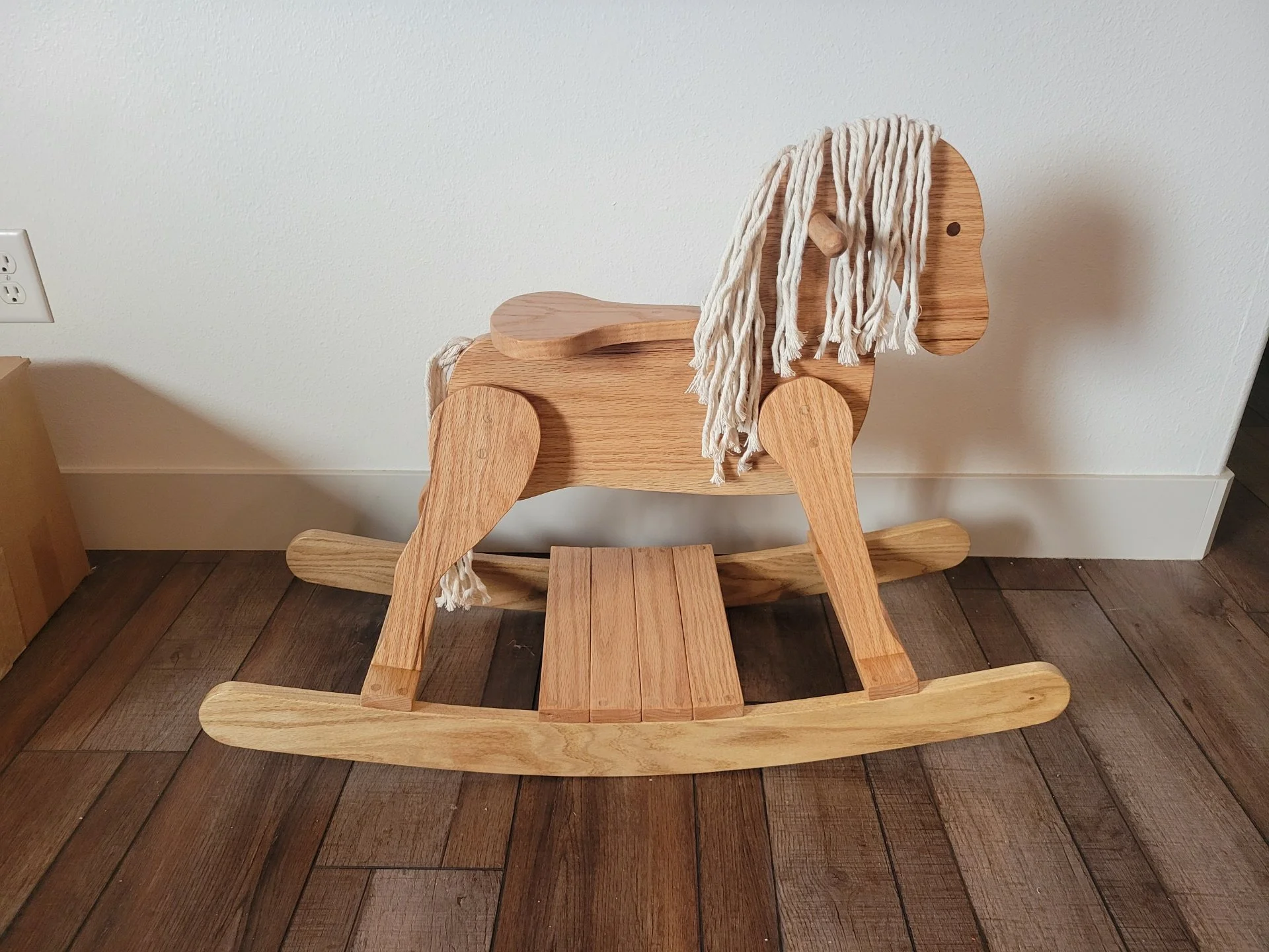 Wooden rocking horse with a white yarn mane, placed against a white wall on a hardwood floor.