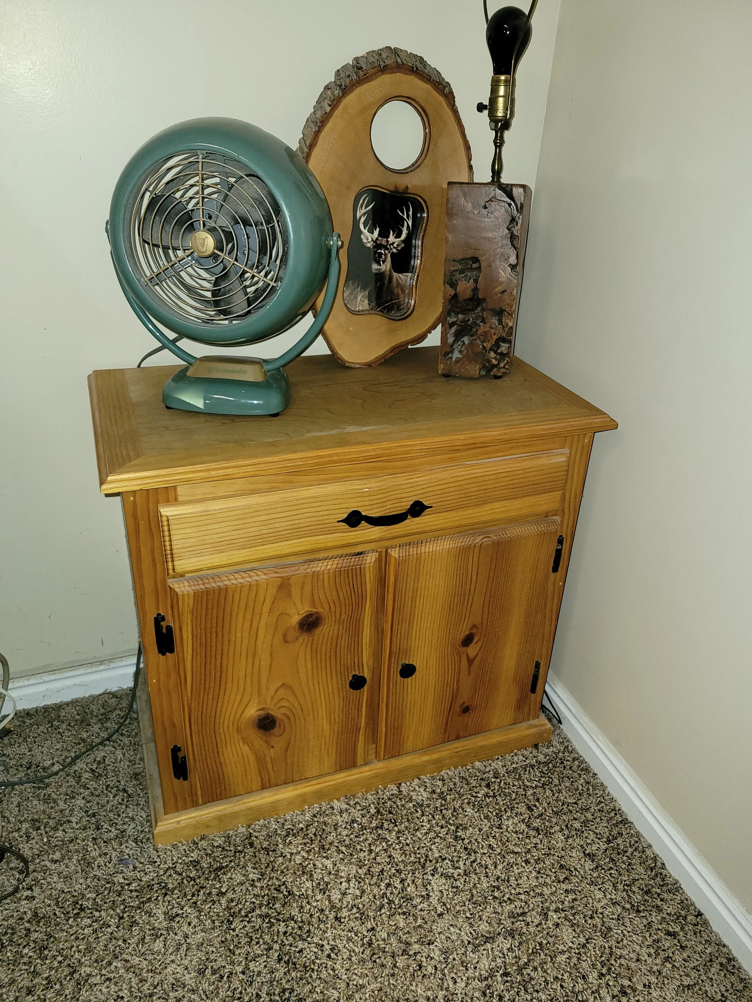 Wooden cabinet topped with a vintage teal fan, a mirror with a deer silhouette, a decorative wood piece, and a black lamp with no shade.