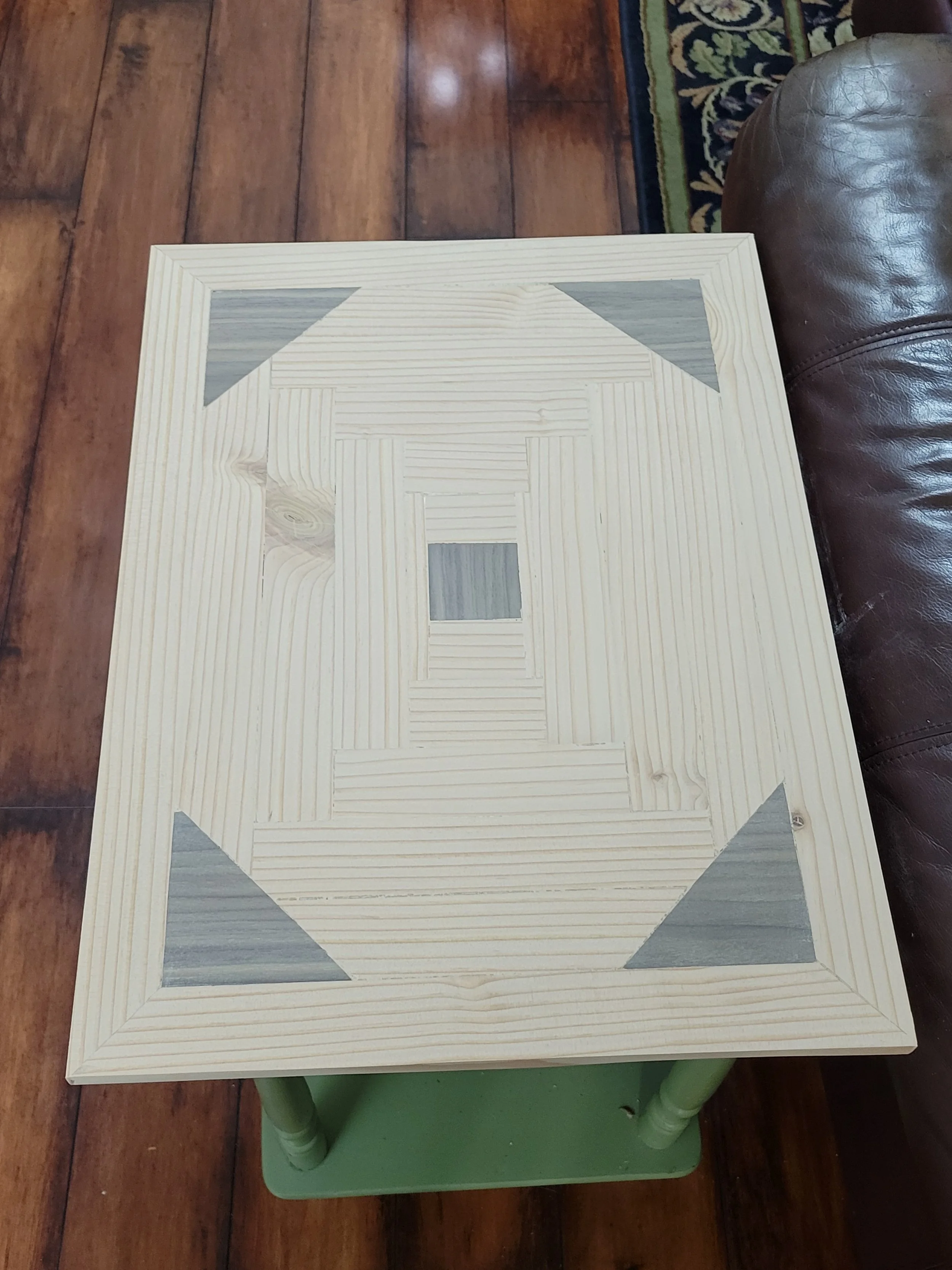 A wooden square table with a decorative pattern of wood grain insets and painted corner triangles, on a hardwood floor next to a leather couch.