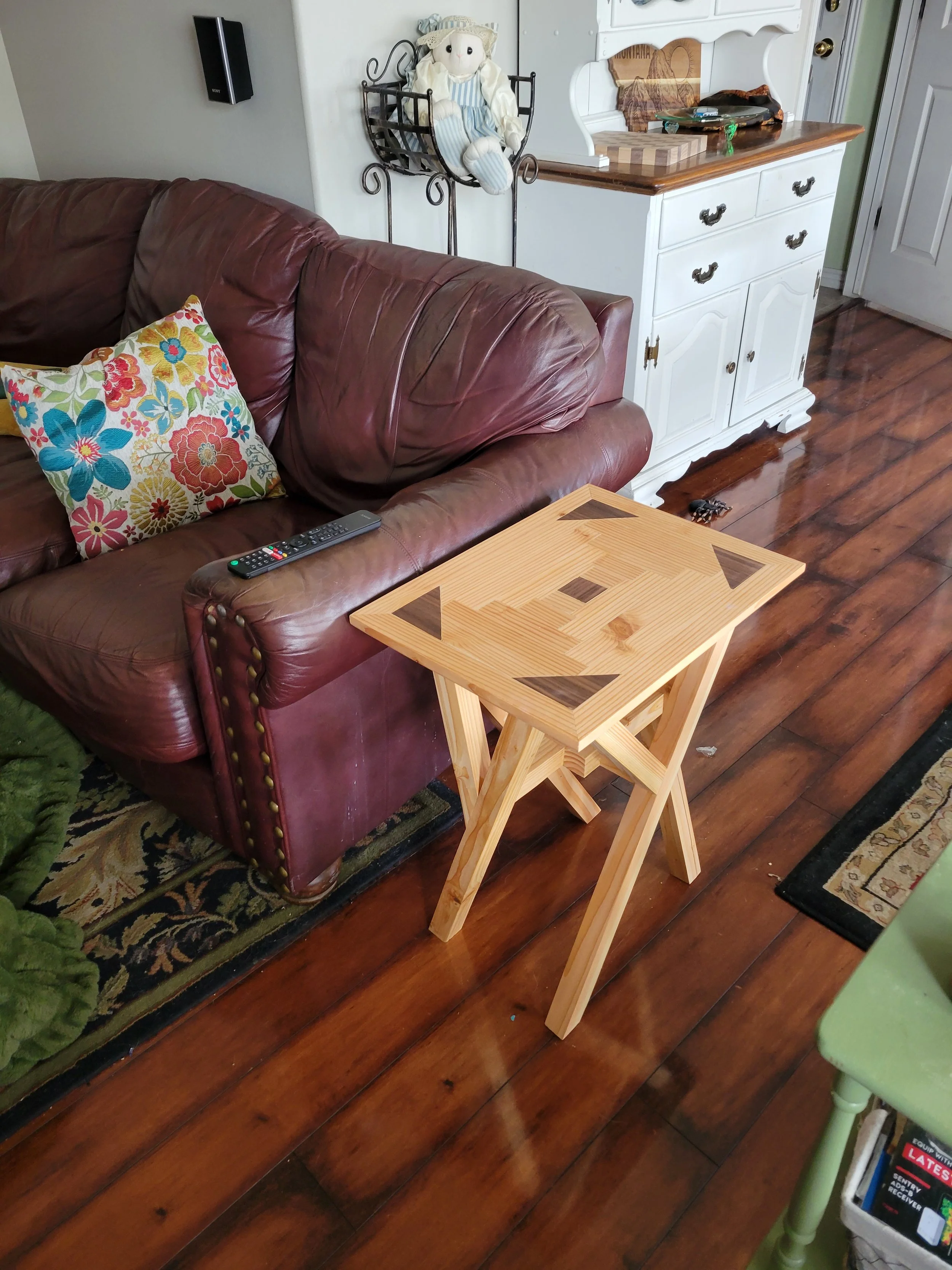 A wooden side table with a geometric inlay design next to a brown leather sofa. The sofa has a colorful floral cushion and a remote control on it. The room has wood flooring, a white dresser with items on top, and a wall-mounted speaker.