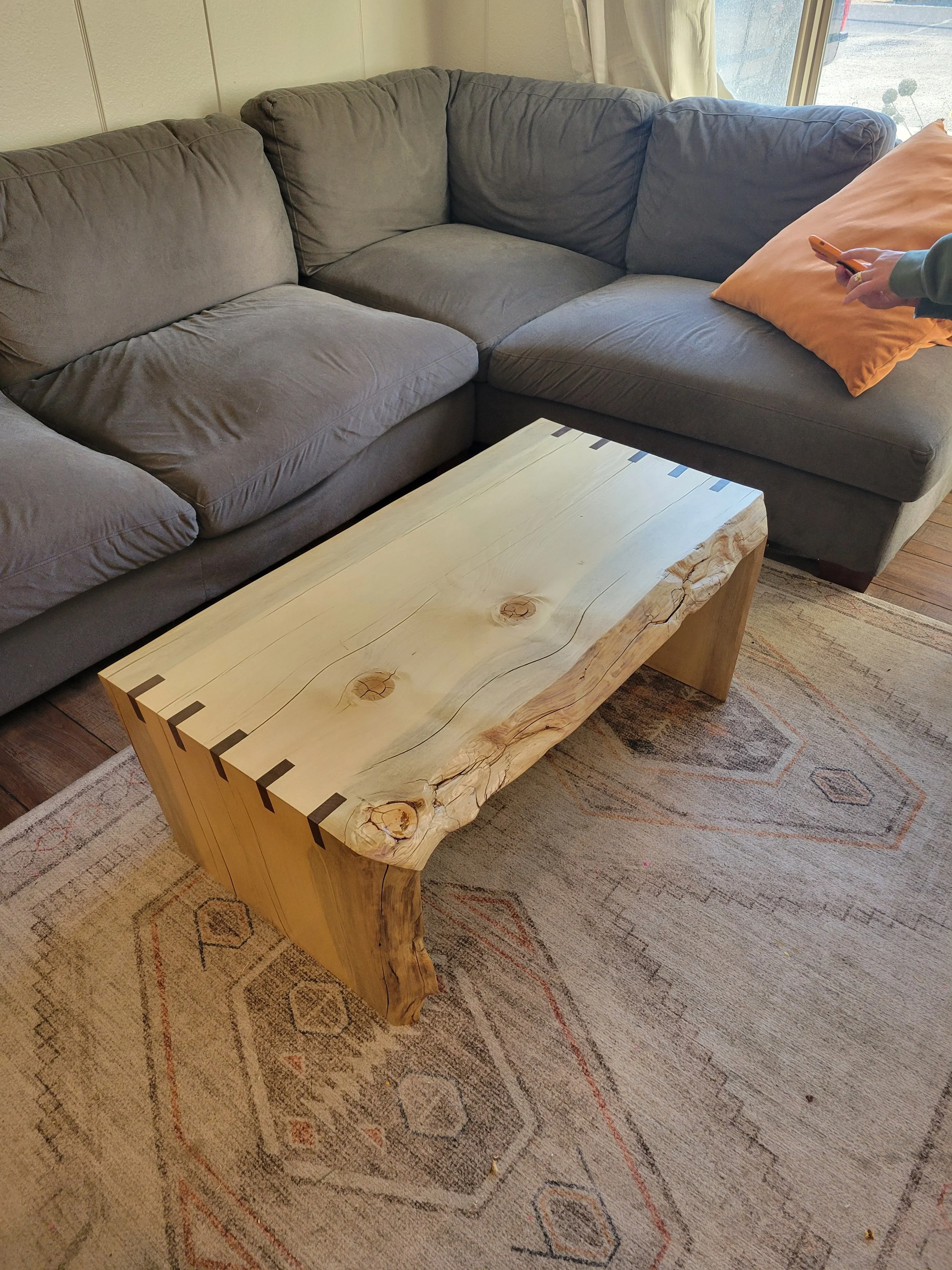 A wooden coffee table with visible knots and a natural edge, placed on a patterned area rug in front of a gray sectional sofa in a living room.